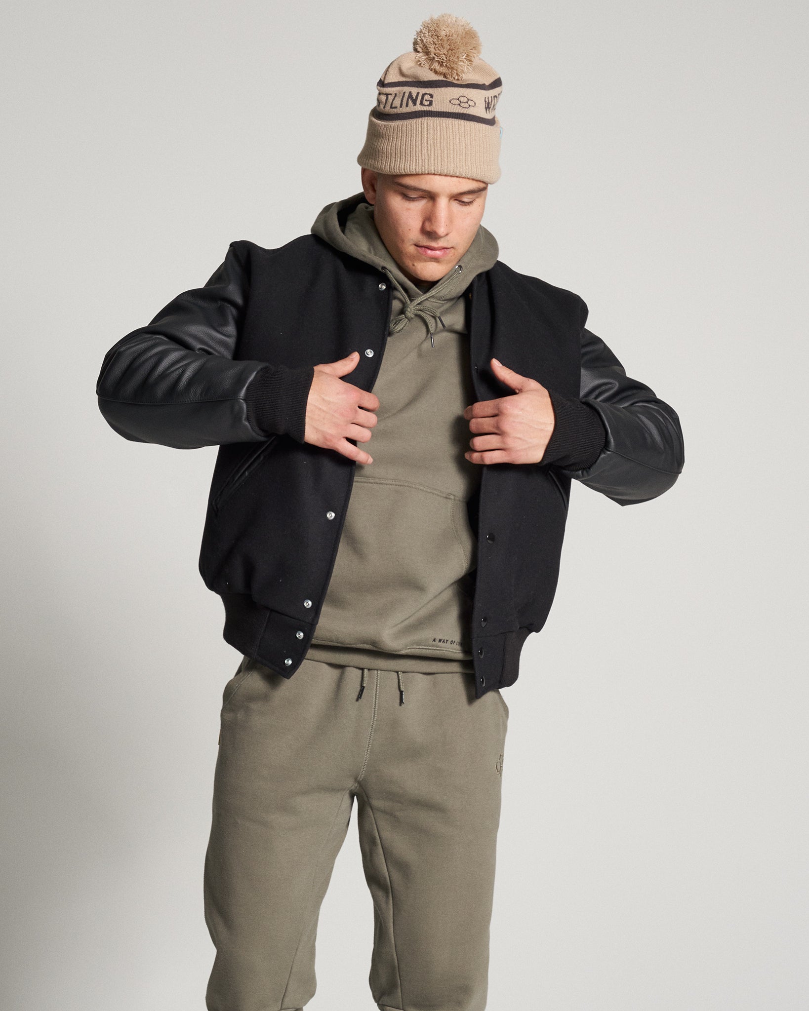 A young man in an urban outfit featuring a black varsity jacket beige beanie and olive green hoodie with matching sweatpants