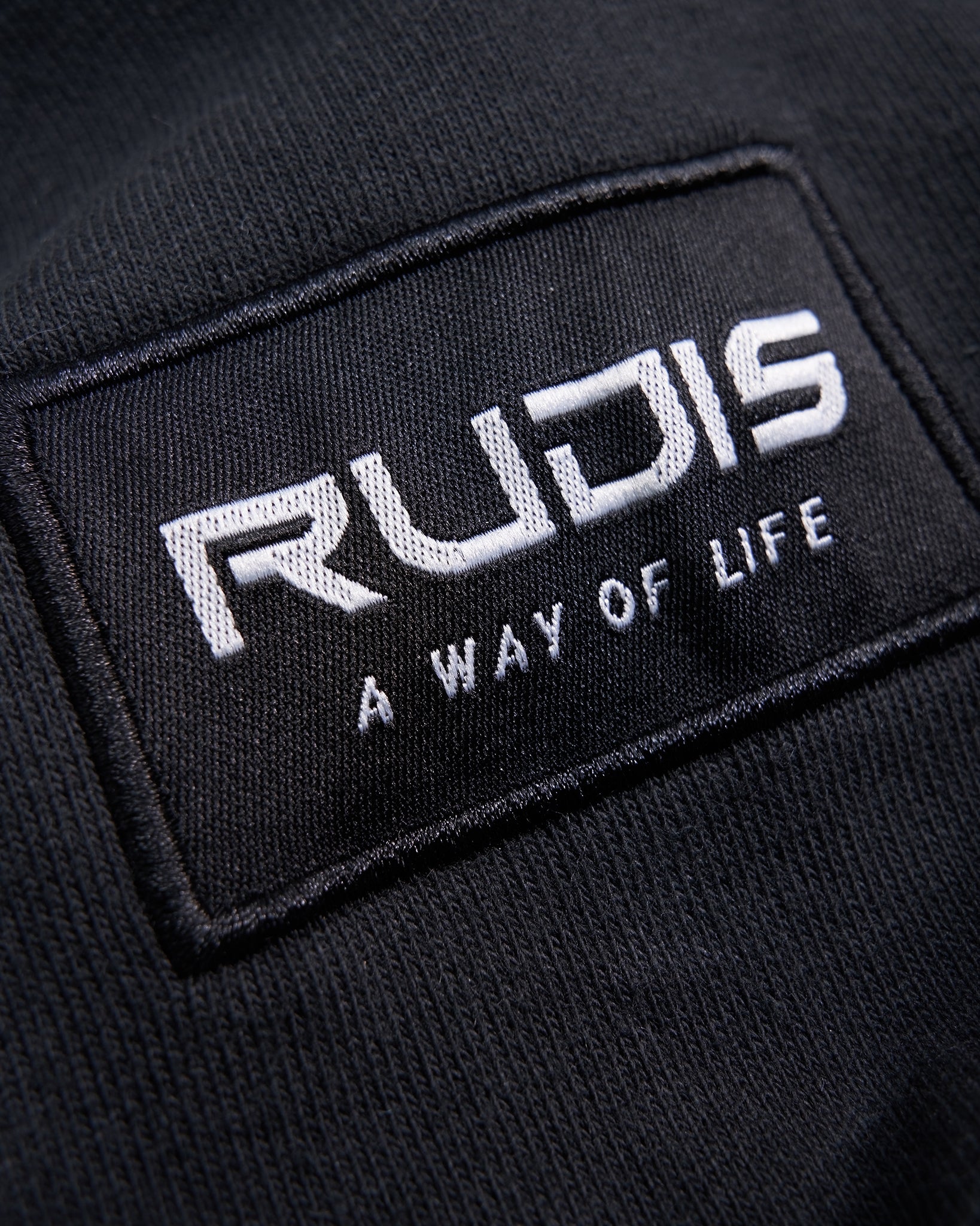 A detailed close-up of a fabric label featuring the word RUDIS and the phrase A WAY OF LIFE in white stitching on a black background