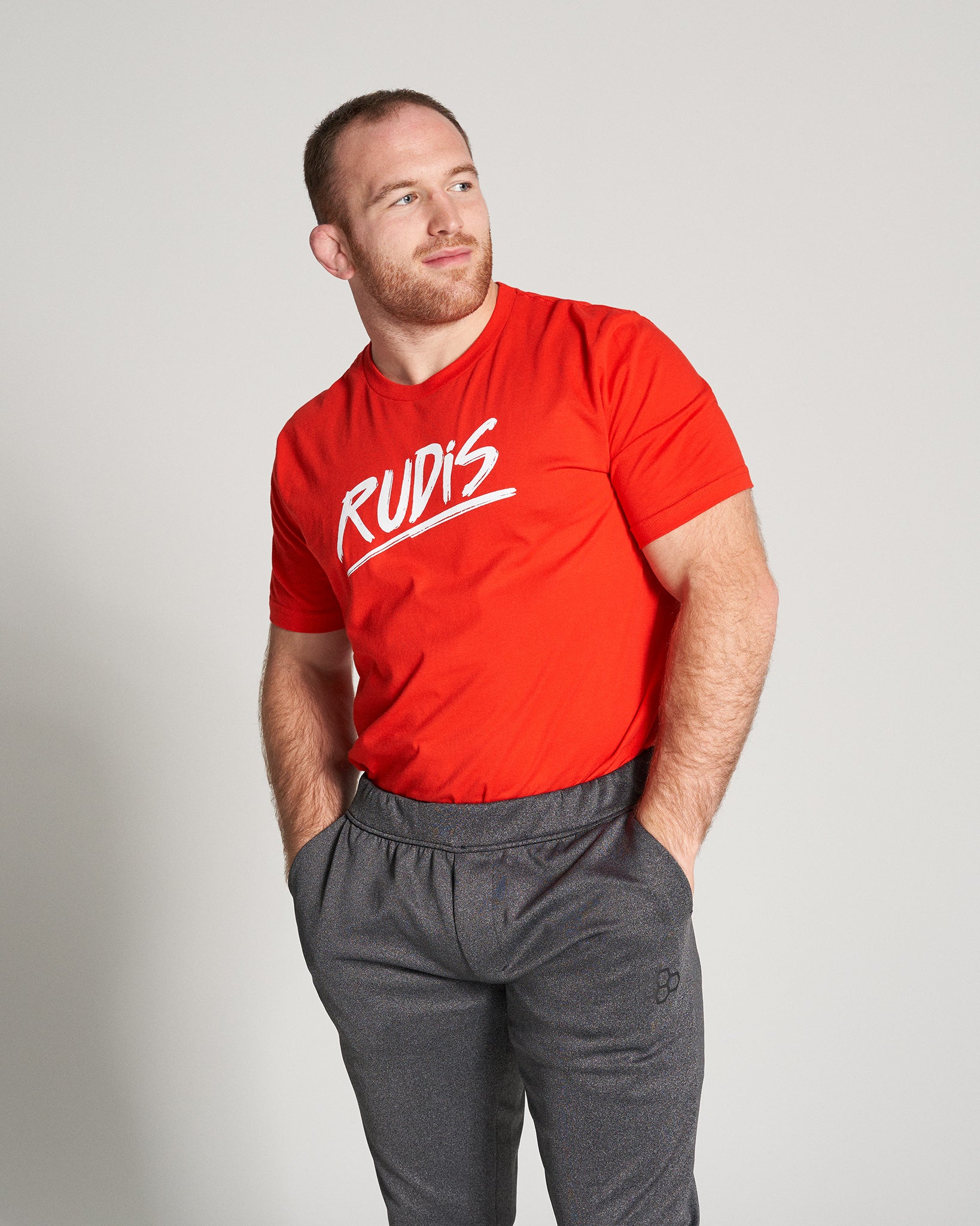 A man with a beard is wearing a bright red t-shirt and gray athletic pants standing confidently with his hands in his pockets showcasing a casual yet stylish look