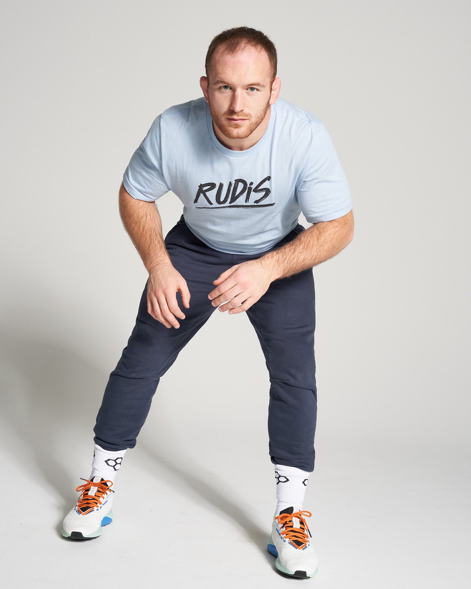 A focused athlete in a light blue t-shirt with RUDIS printed on it wearing dark pants and stylish sneakers demonstrating an athletic stance on a light gray background