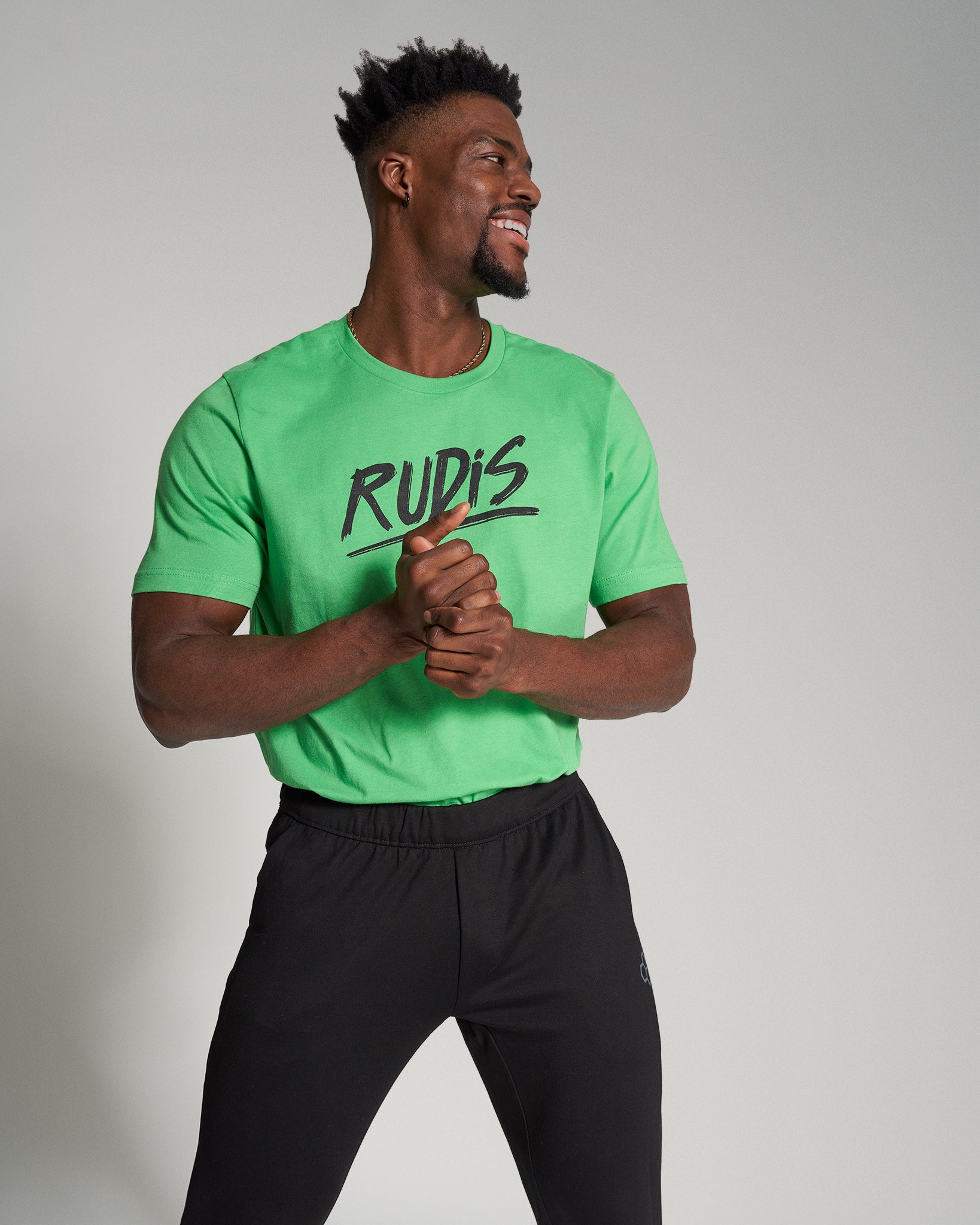 A smiling man in a vibrant green t-shirt with RUDIS printed on it paired with black athletic pants exudes energy and confidence