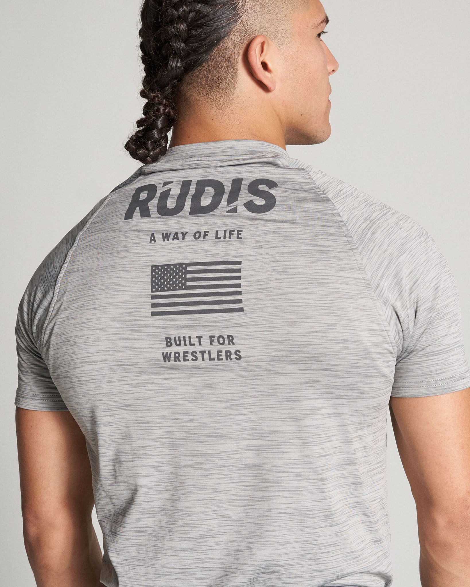 A man is seen wearing a gray athletic shirt featuring the word RUDIS and a small American flag on the back emphasizing a wrestling theme