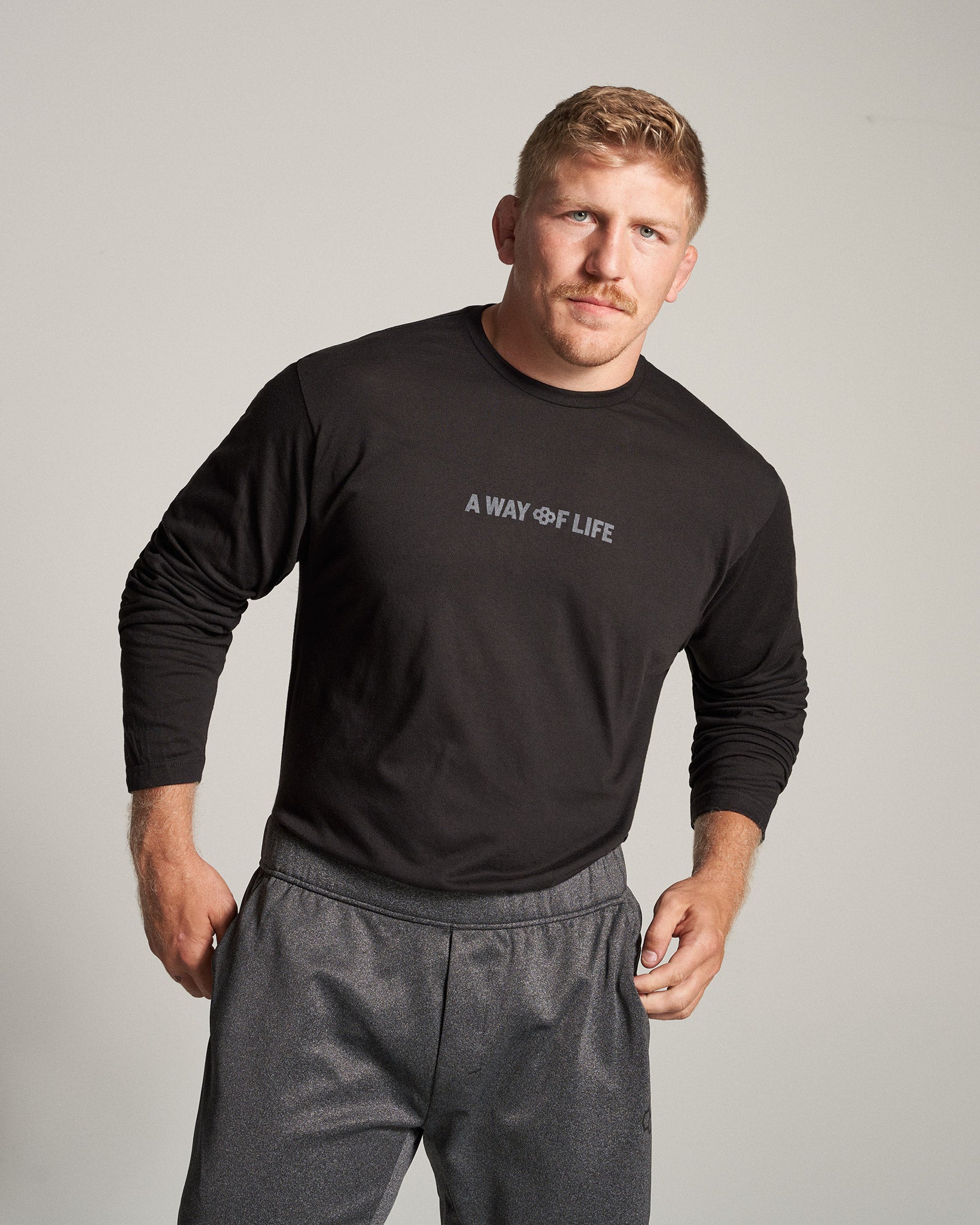 A muscular man dressed in a long-sleeve black shirt and gray athletic pants standing confidently with a slight smile against a neutral background