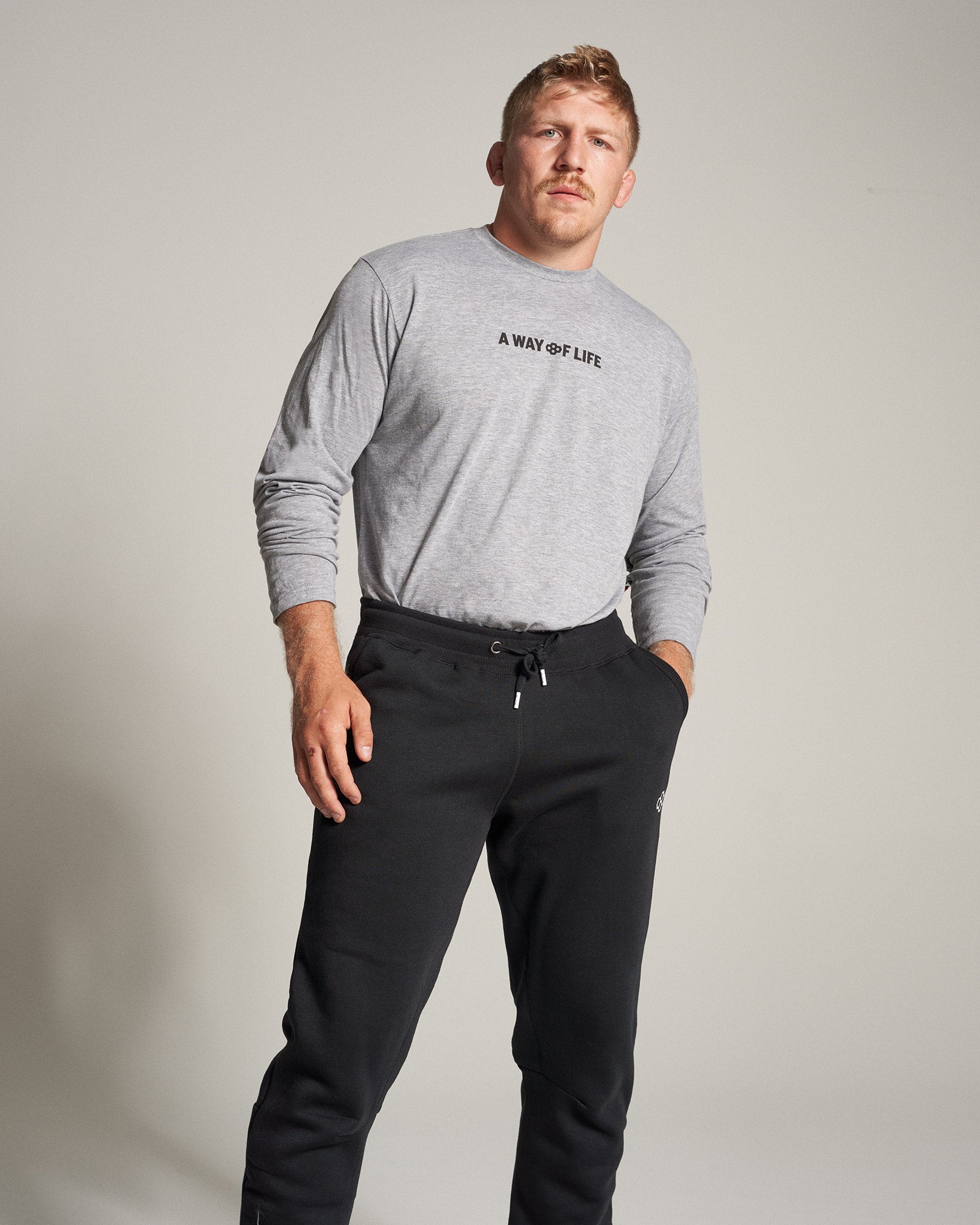 A man stands confidently wearing a gray long-sleeve shirt that reads A WAY OF LIFE and black sweatpants showcasing a relaxed yet stylish athletic look