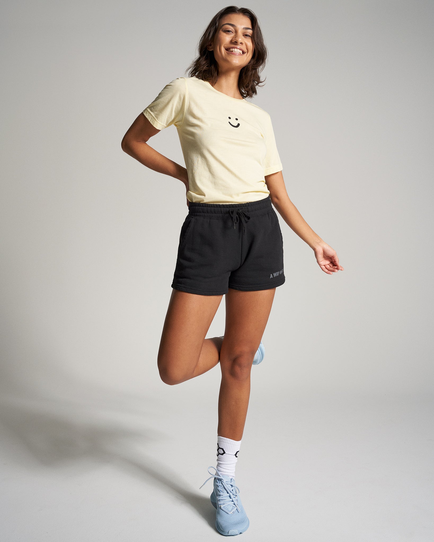 A young woman is playfully posing in a casual outfit consisting of a light yellow smiley face t-shirt and black shorts complemented by stylish blue sneakers and patterned socks