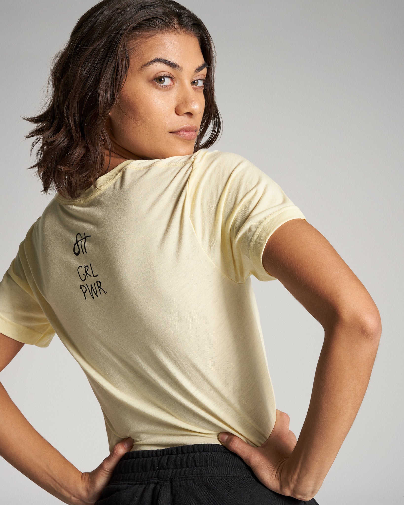 A woman poses confidently in a light yellow shirt with the words GRL PWR printed on the back exuding empowerment and style