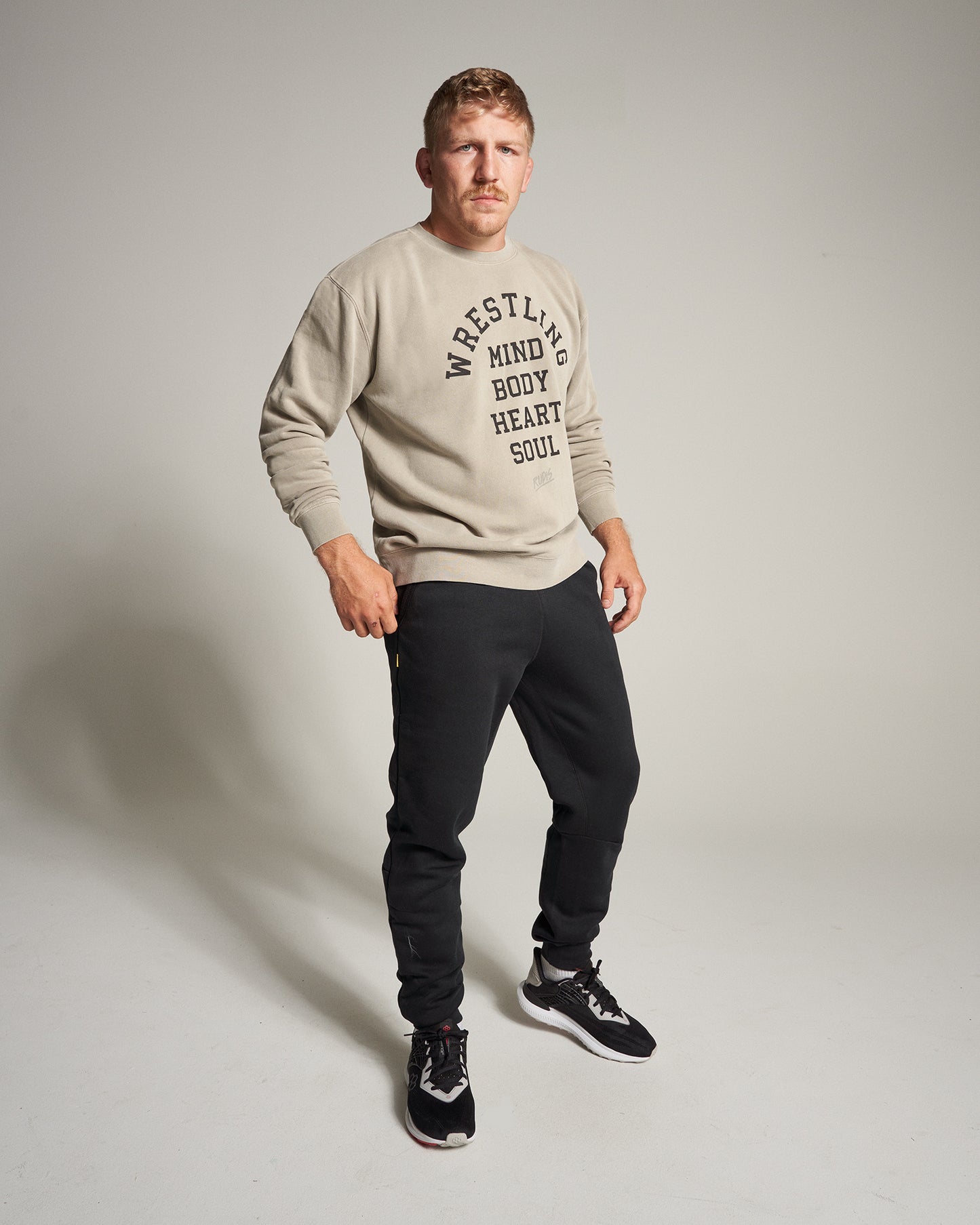 A man stands confidently in a beige sweatshirt that reads WRESTLING MIND BODY HEART SOUL paired with black sweatpants and black athletic shoes set against a neutral background