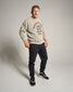 A man stands confidently in a beige sweatshirt that reads WRESTLING MIND BODY HEART SOUL paired with black sweatpants and black athletic shoes set against a neutral background