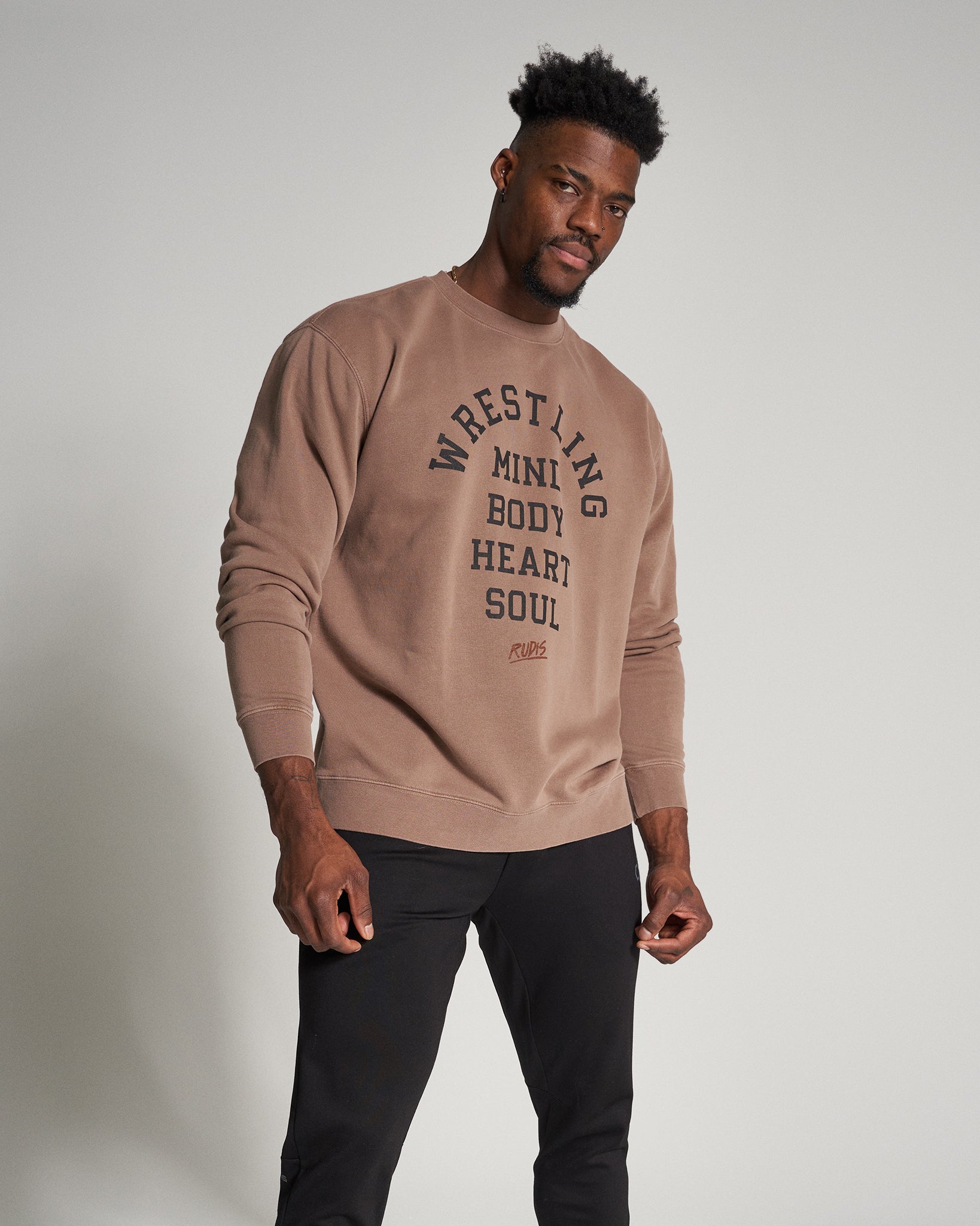 A man wears a brown sweatshirt featuring bold black text that reads WRESTLING MINI BODY HEART SOUL paired with black pants in a minimalist studio setting