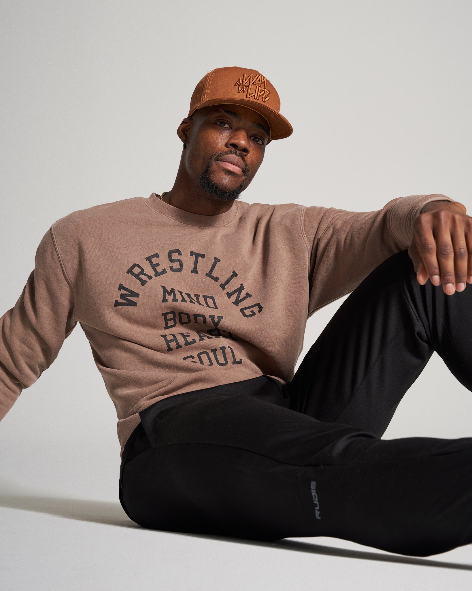 A man sits casually wearing a brown sweatshirt with a bold black print and black pants complemented by a tan cap exuding a relaxed streetwear style