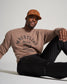 A man sits casually wearing a brown sweatshirt with a bold black print and black pants complemented by a tan cap exuding a relaxed streetwear style