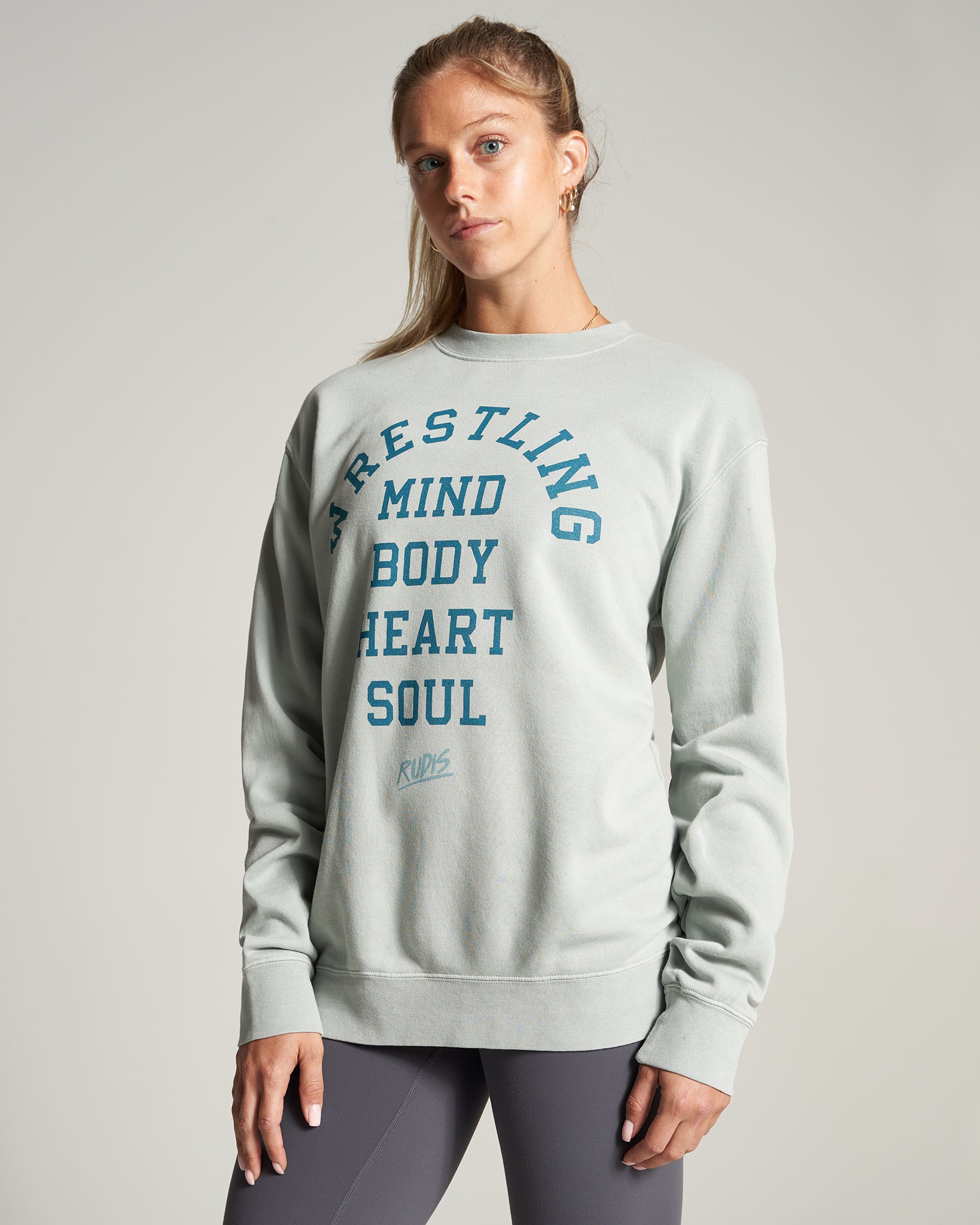 A woman poses in a light green sweatshirt featuring bold text that reads WRESTLING MIND BODY HEART SOUL paired with dark leggings conveying a sporty and motivational aesthetic