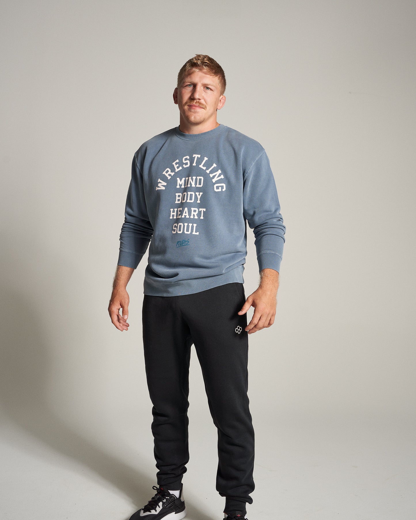 A man poses confidently in a blue sweatshirt and black sweatpants featuring motivational text related to wrestling