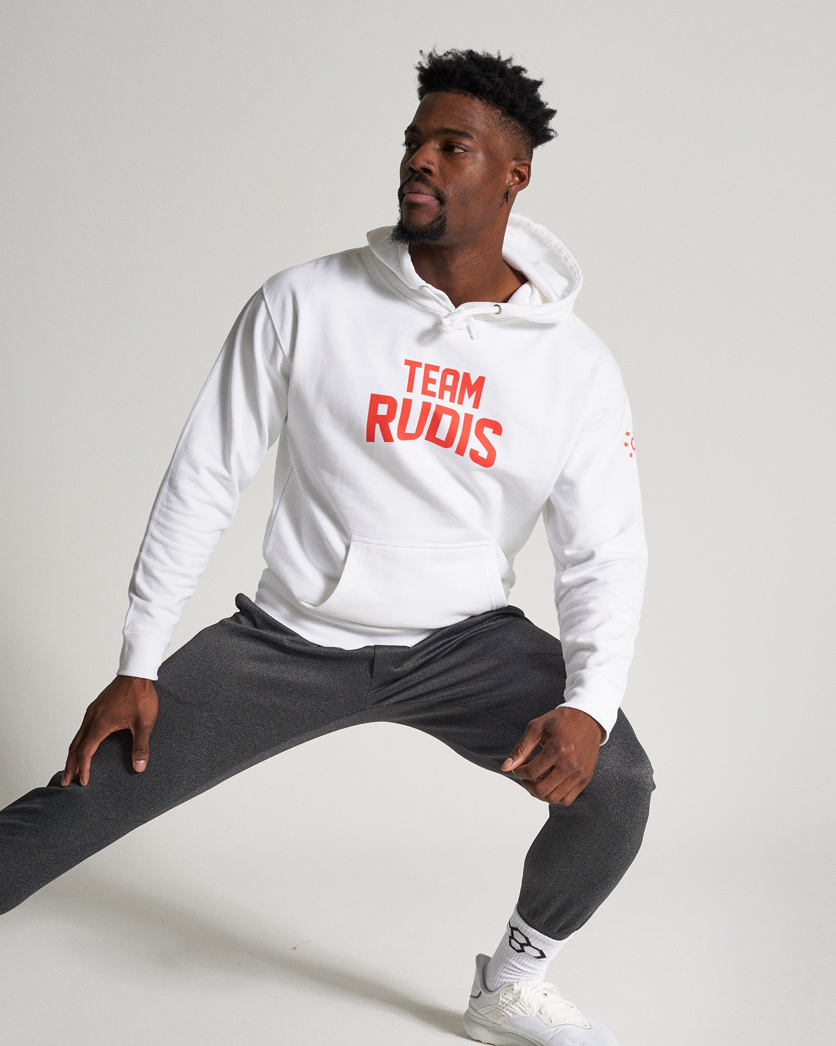 A man performing a lunge in athletic wear featuring a white hoodie with TEAM RUDIS printed in bold red and dark gray joggers