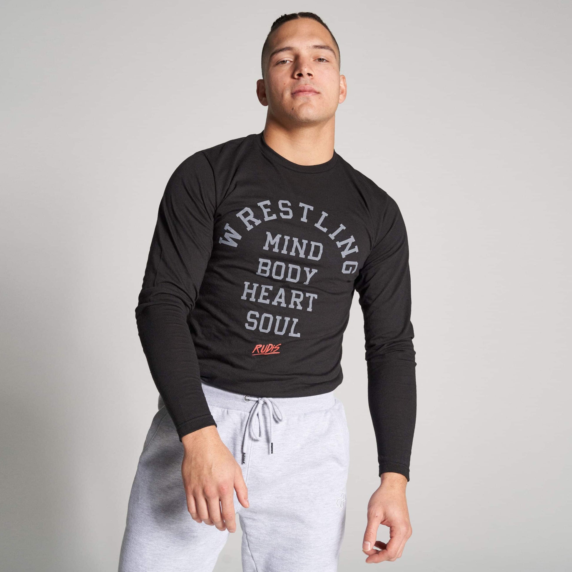 A young male athlete poses confidently in a long-sleeve black shirt with motivational wrestling-themed text and comfortable gray sweatpants