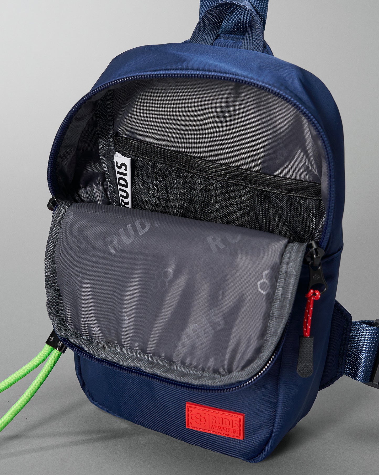 A navy blue sling bag with a gray padded flap, black mesh pocket, and a bold red logo patch combines functionality with style.