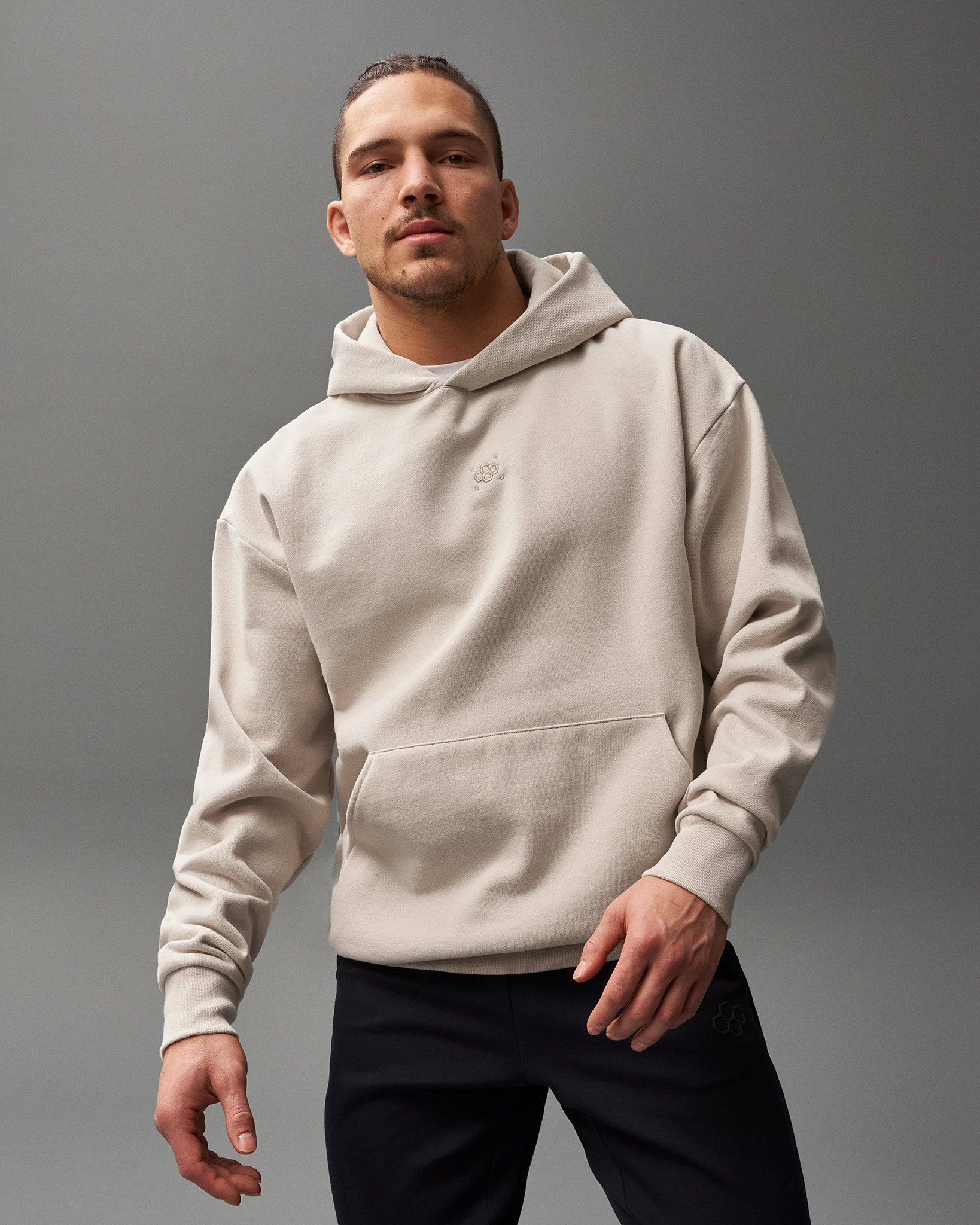 A man confidently poses in a light beige hoodie with a front pocket and embroidered logo, paired with black pants against a solid gray background.