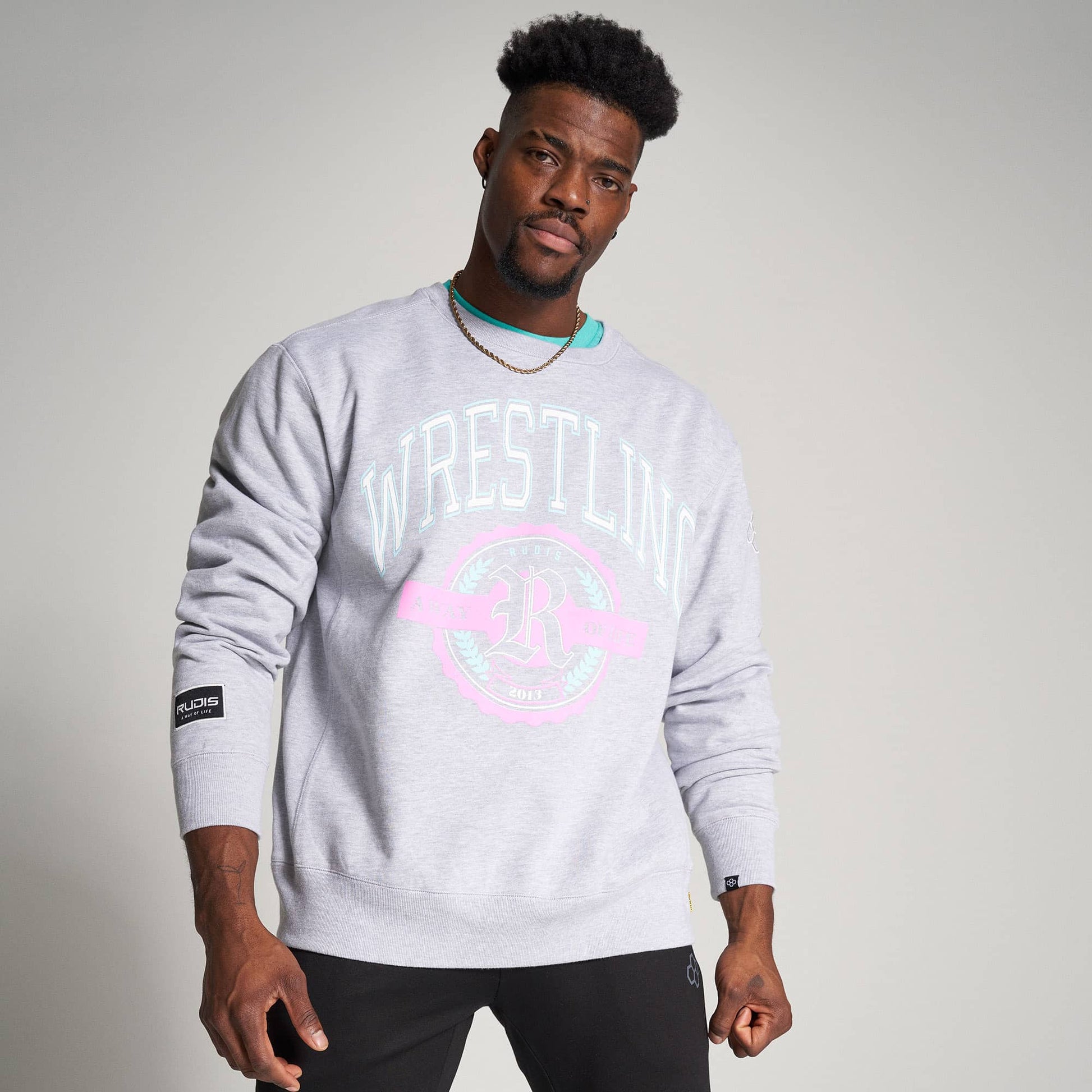 A male model poses confidently wearing a light gray sweatshirt with a prominent wrestling-themed design and black pants