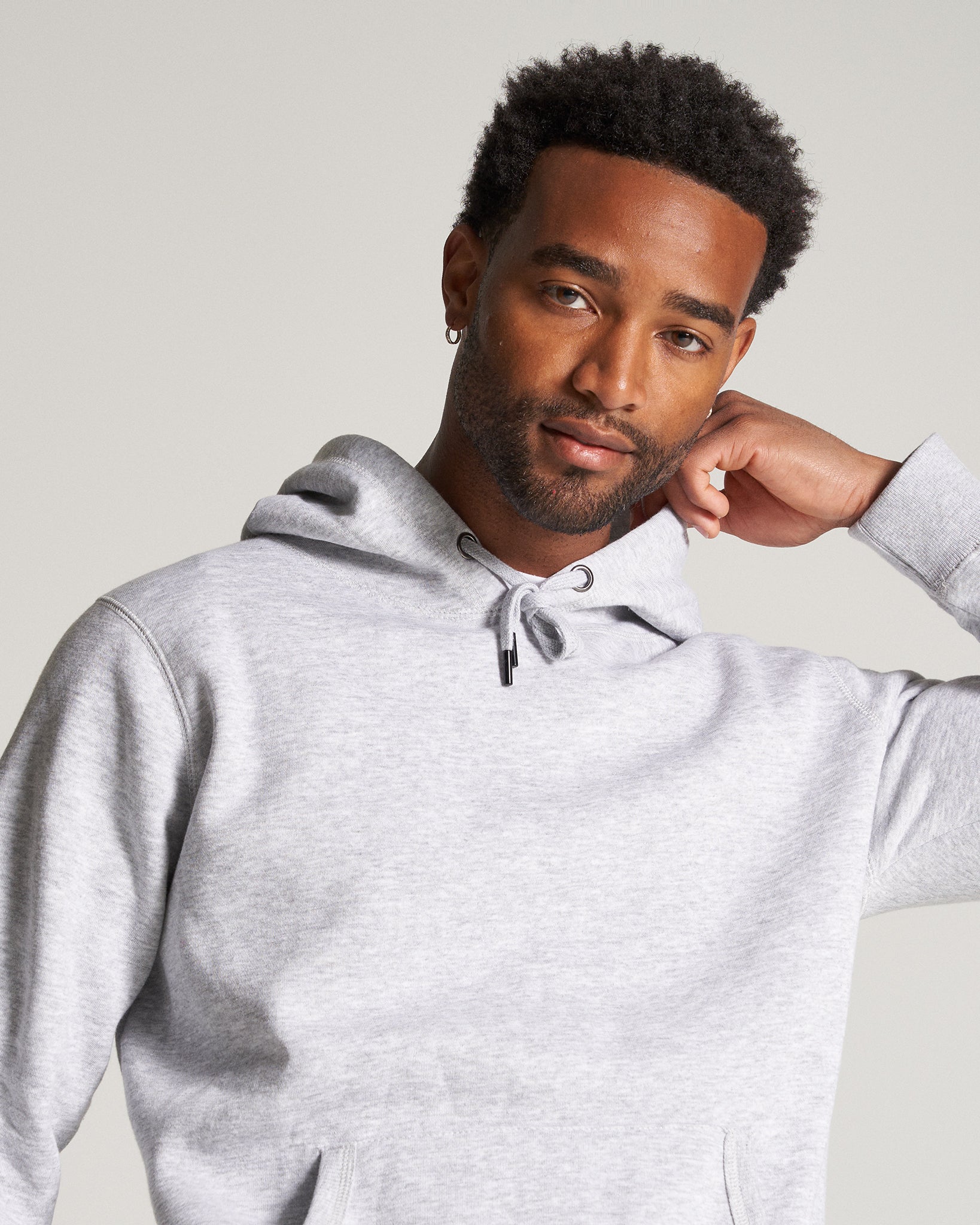 A fashionable black man confidently poses in a light gray hoodie showcasing a casual yet stylish look complemented by his neat haircut and subtle earring