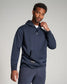 A man wearing a navy blue hoodie with a front pocket casually adjusting his collar in a softly lit background