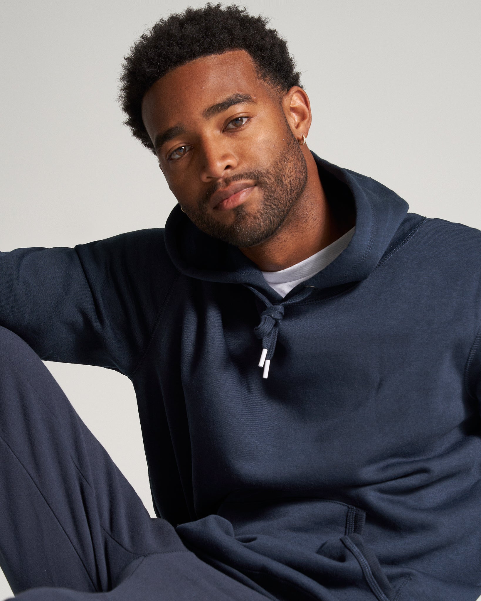 A young man is shown wearing a dark navy hoodie with a relaxed expression seated in a casual pose against a light gray background