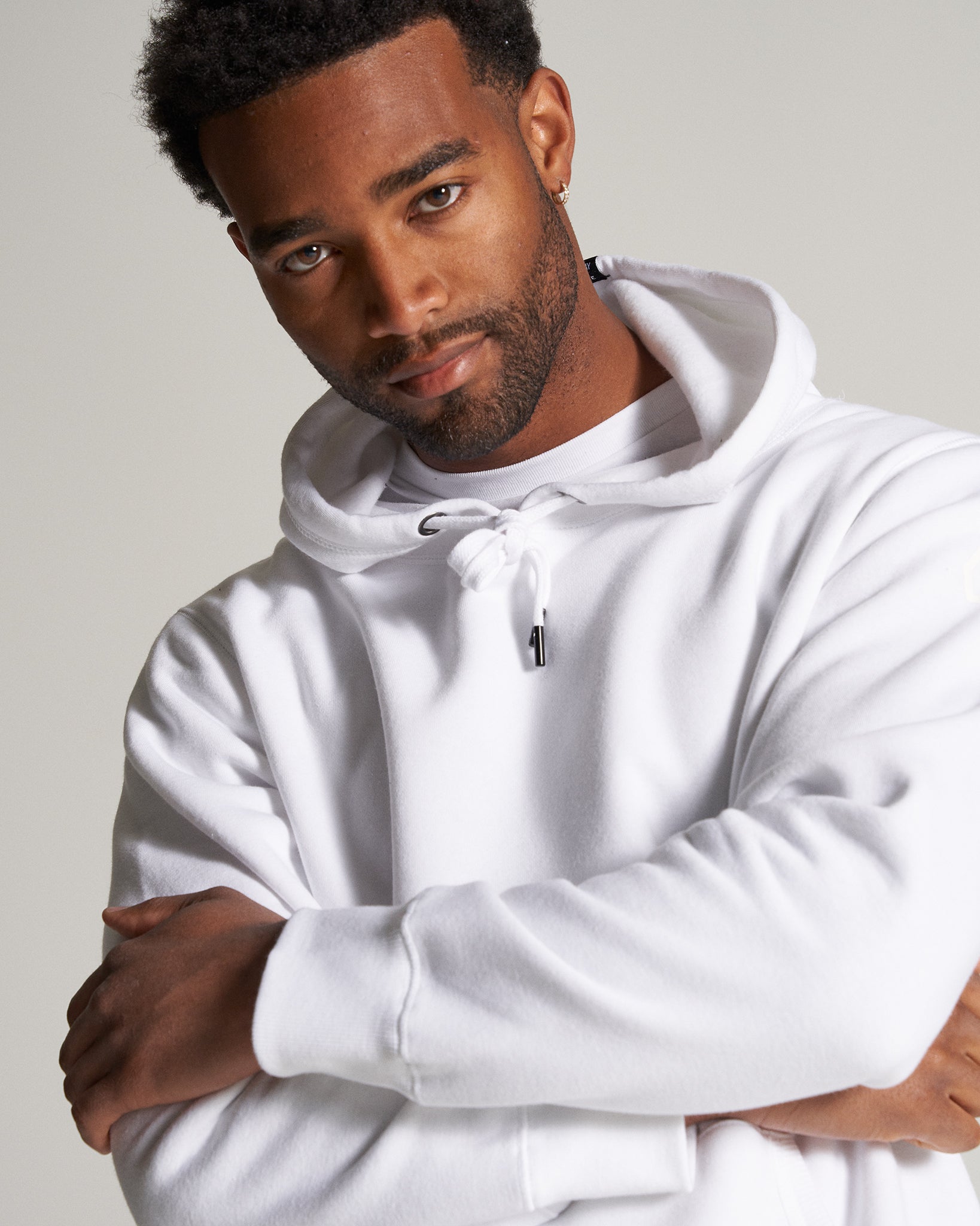 A young man with a neatly groomed beard is wearing a white hoodie looking confidently at the camera with his arms crossed