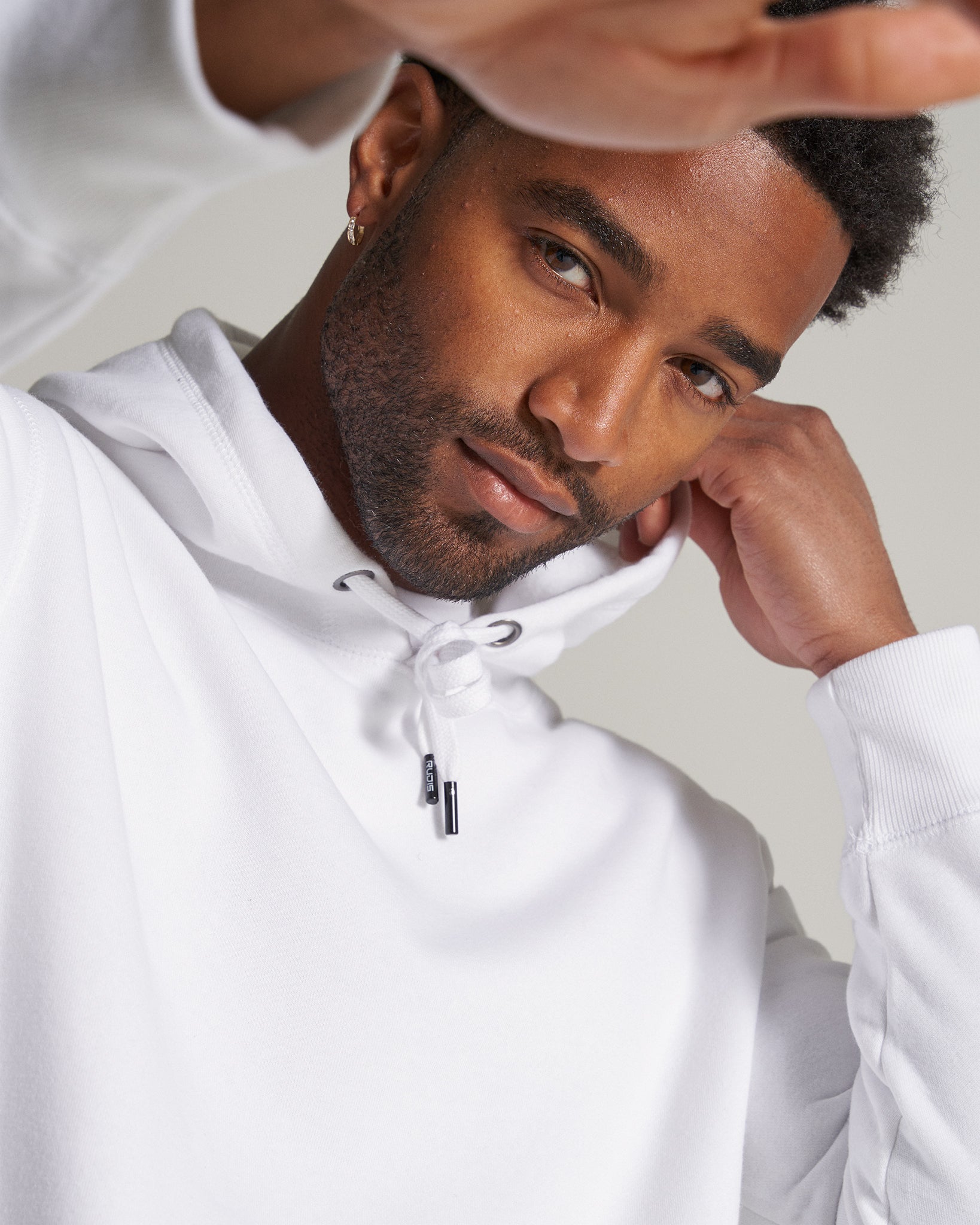 A close-up of a smiling man wearing a white hoodie looking confidently at the camera while partially raising his hand in a relaxed pose