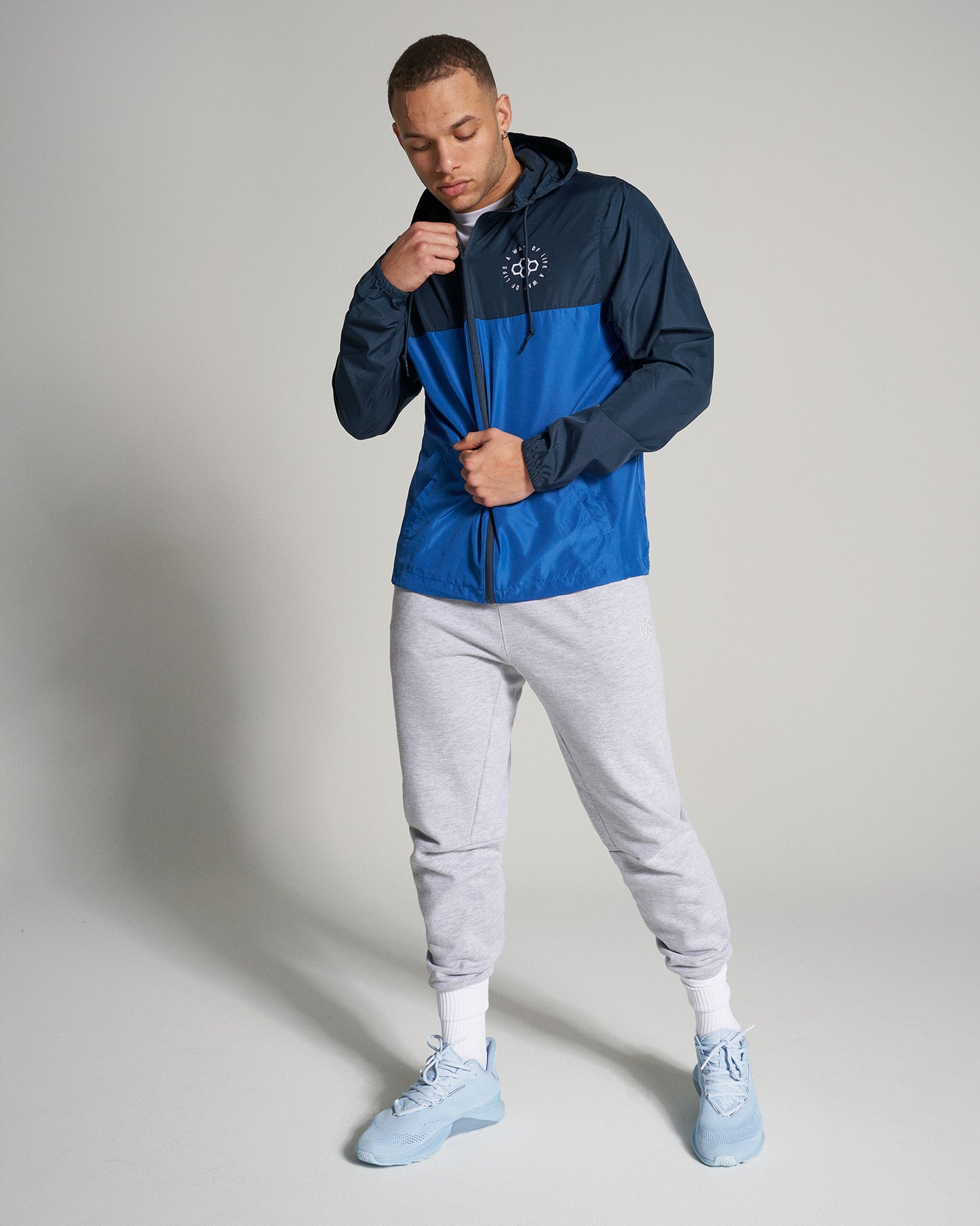 A young man is wearing a blue and navy windbreaker jacket paired with gray sweatpants and light blue sneakers looking stylish and relaxed against a neutral background