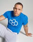 A man with short hair and blue eyes poses in a vibrant blue t-shirt featuring a geometric honeycomb logo paired with light gray sweatpants creating a casual yet stylish look against a neutral background