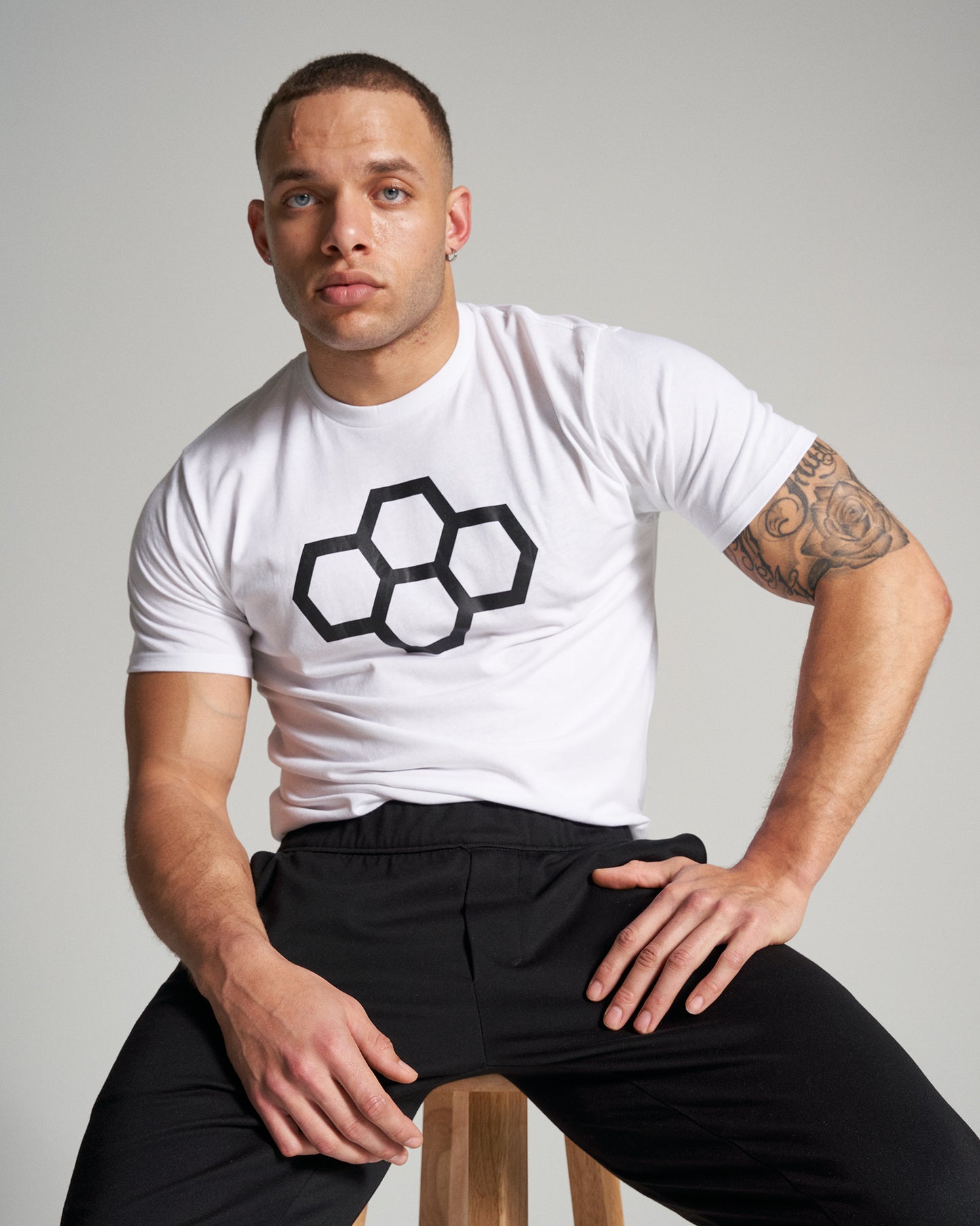 A muscular man seated on a wooden stool wearing a white t-shirt with a black hexagonal logo and black pants against a gray background