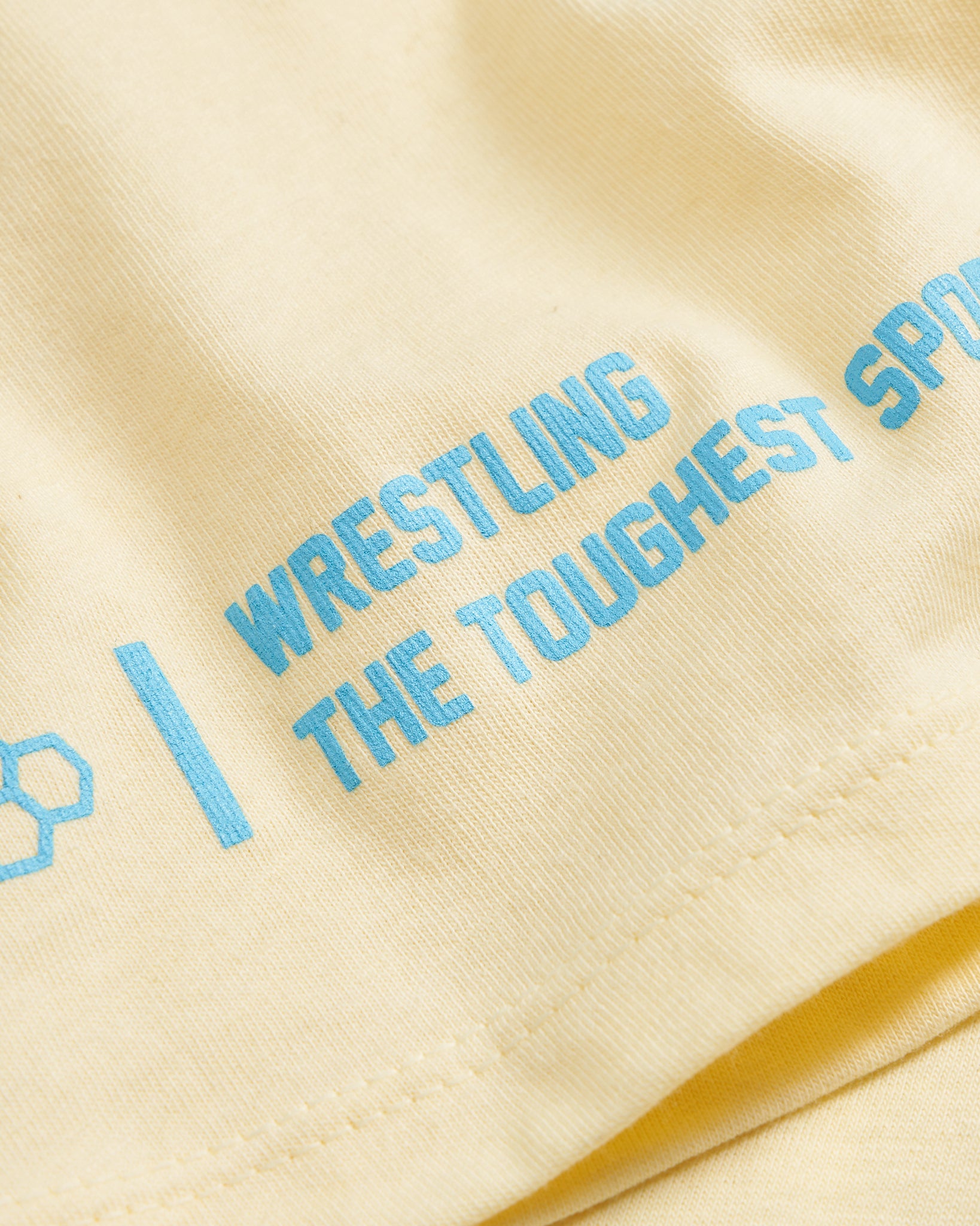 A close-up view of a light yellow fabric featuring bold blue text that reads WRESTLING THE TOUGHEST SPORT