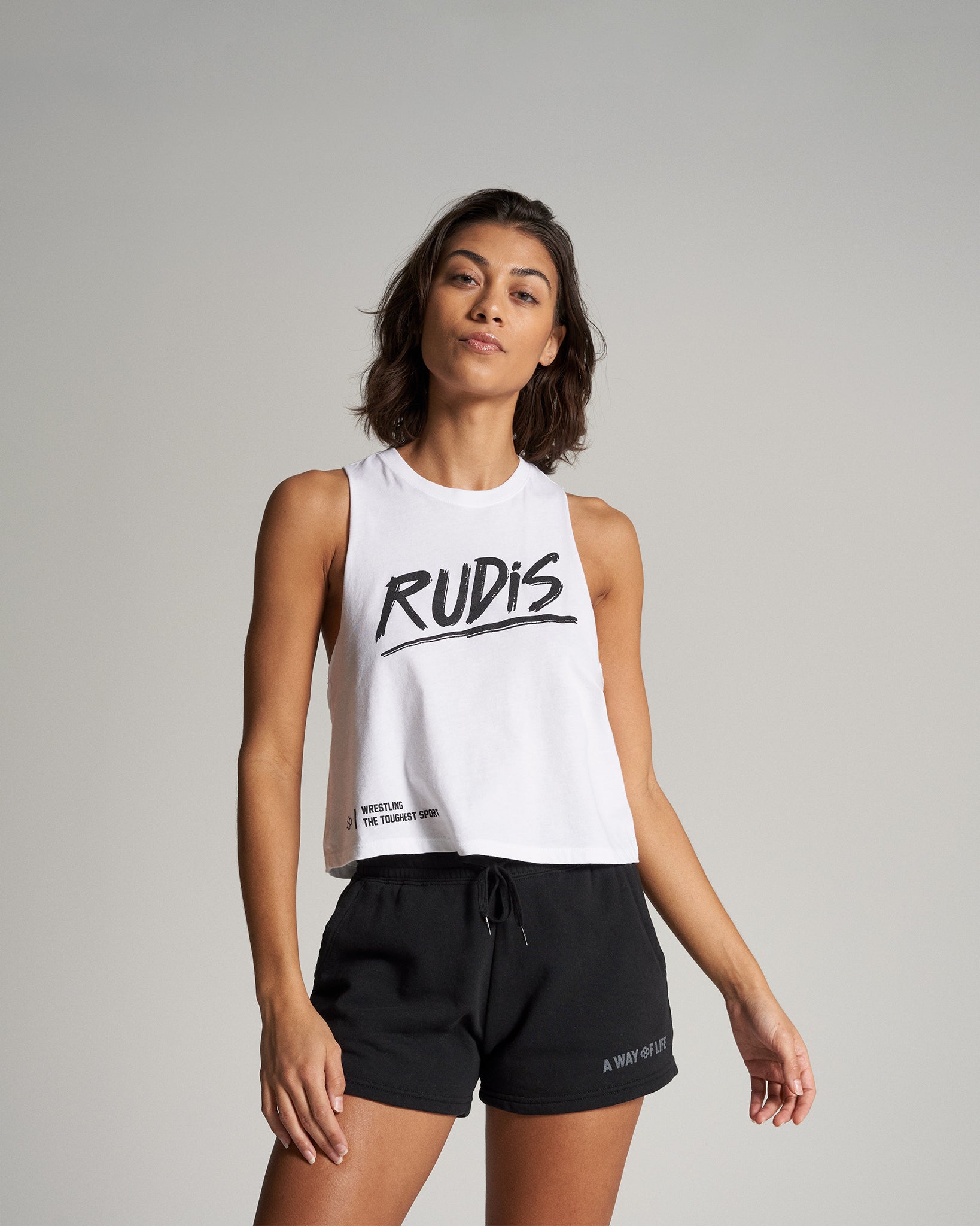 A model wears a stylish white crop top with the word RUDIS in bold black letters paired with black athletic shorts staged against a neutral background