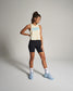 A woman stands in an athletic outfit featuring a cropped beige tank top with blue lettering and black shorts complemented by light blue sneakers and patterned socks