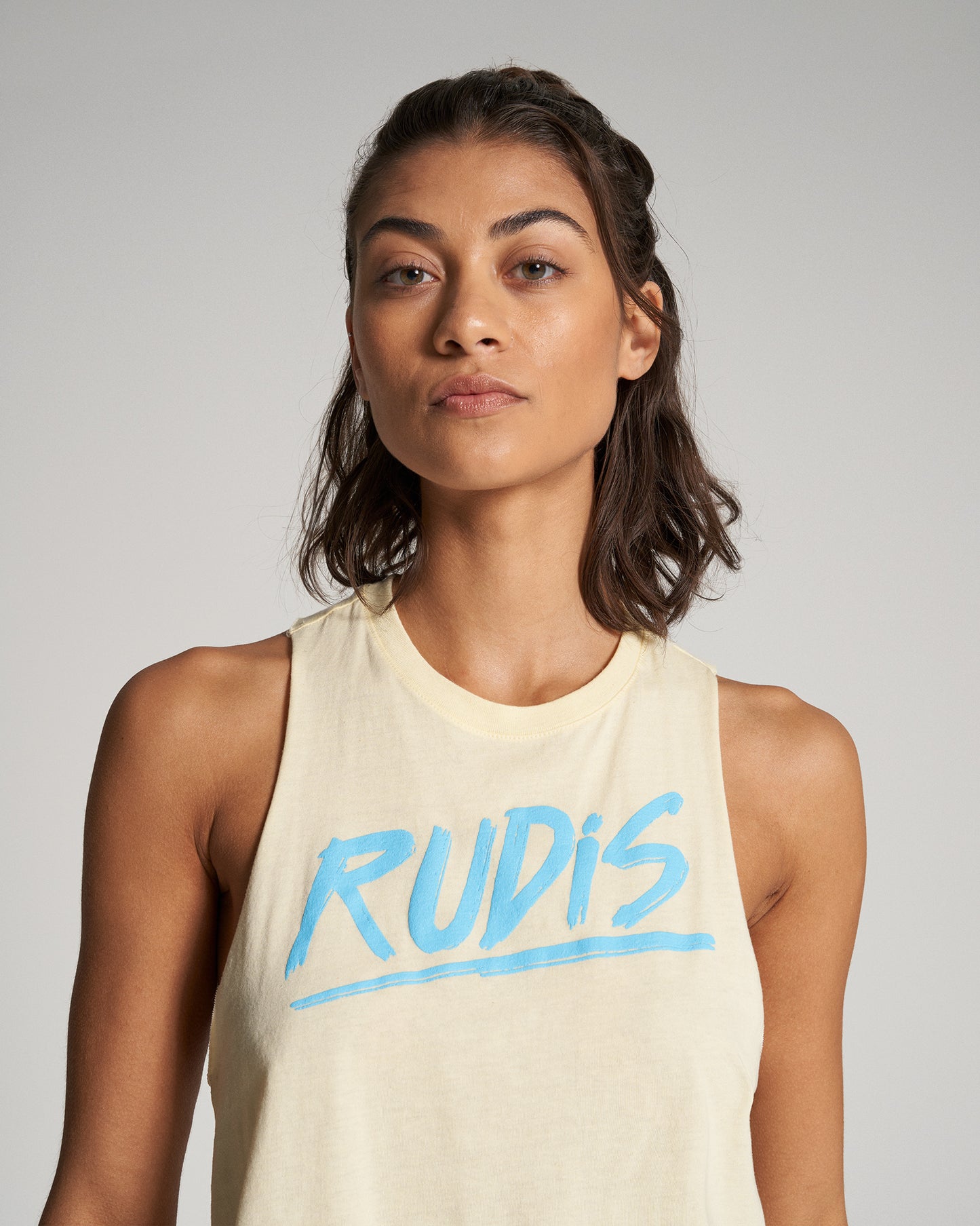 A young woman models a light yellow sleeveless shirt with RUDIS printed in bold blue letters against a neutral gray background