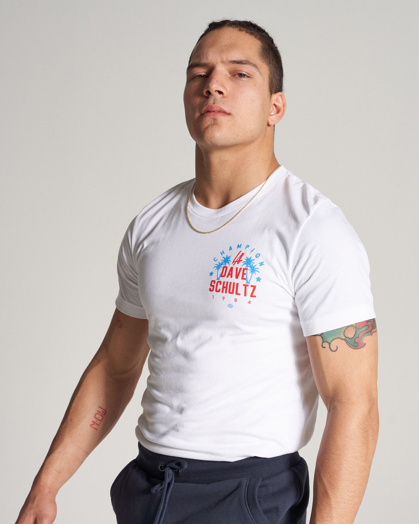 A confident young man poses in a fitted white t-shirt featuring colorful graphics showcasing his muscular build against a neutral background