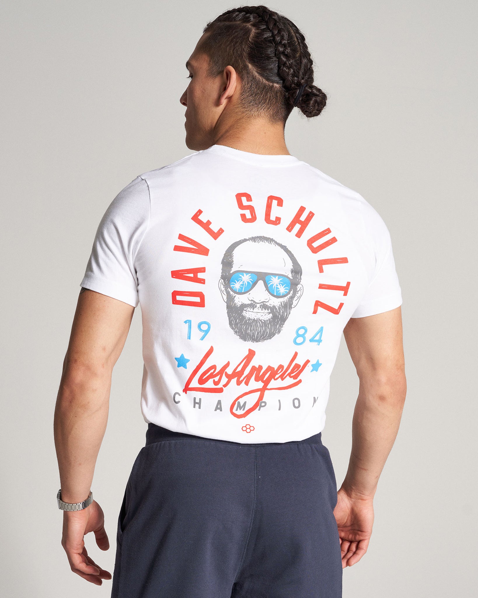 A man wears a white t-shirt featuring a colorful graphic of Dave Schultz on the back including sunglasses bright colors and the text Los Angeles Champion