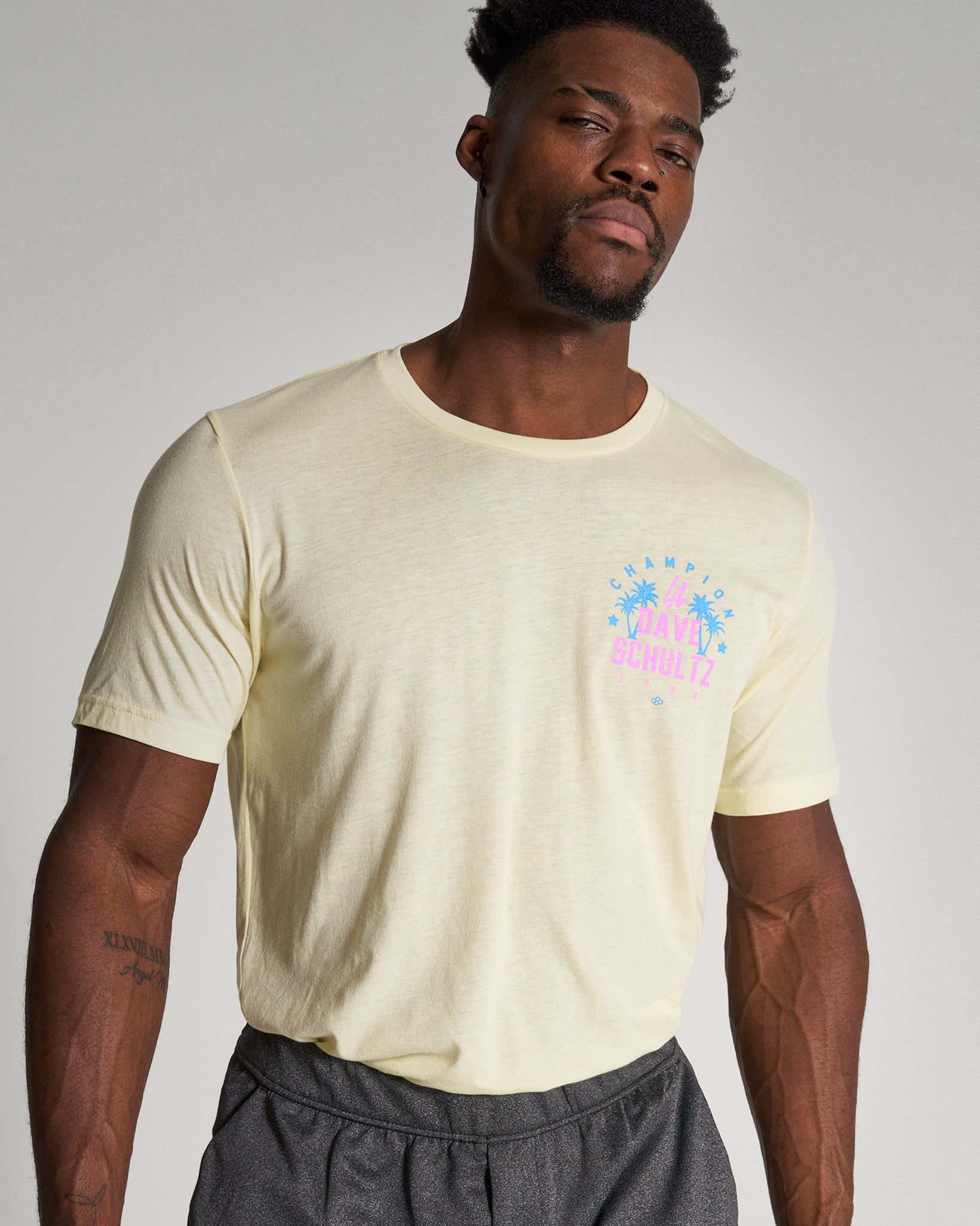 A model displays a light yellow t-shirt featuring a vibrant graphic design with palm trees and a playful text complemented by dark gray shorts creating a casual summer look