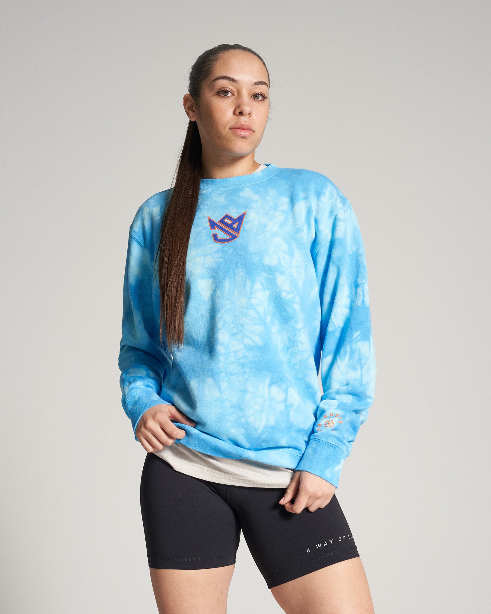 A young woman poses confidently in a blue tie-dye sweatshirt with an emblem paired with black shorts showcasing a casual yet stylish athletic look