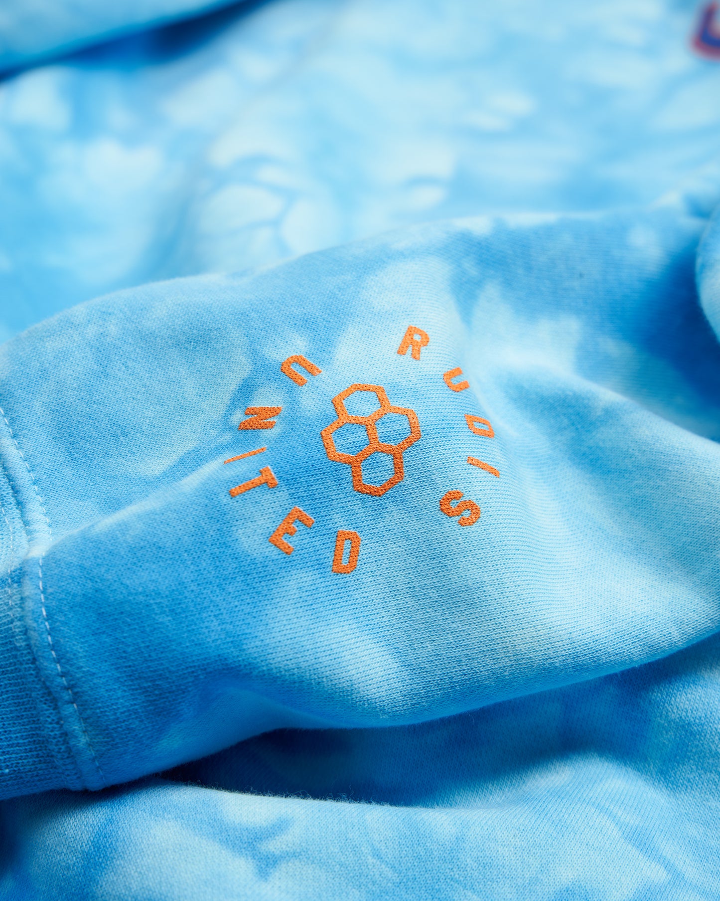 A close-up of a light blue fabric shirt featuring orange text that reads Rudis United along with a honeycomb graphic