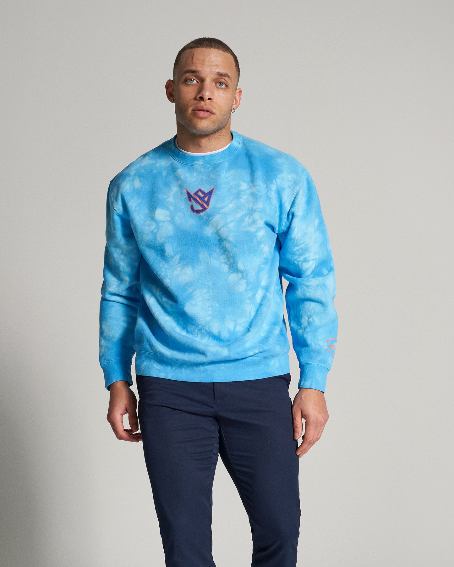 A young man is wearing a vibrant blue tie-dye sweatshirt paired with dark trousers standing against a neutral background