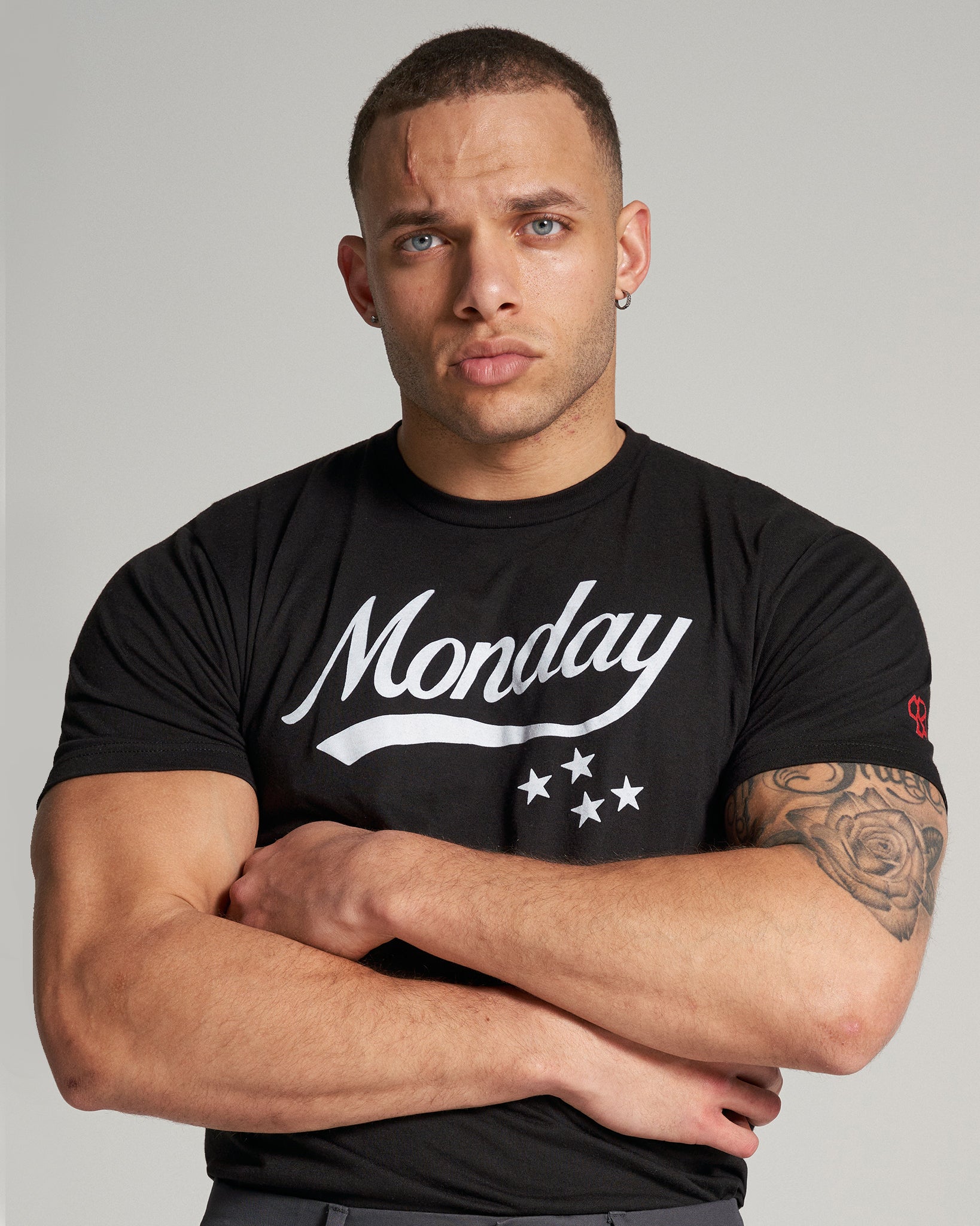 A muscular man with a serious expression stands with his arms crossed wearing a black T-shirt featuring the word Monday in white with decorative stars below it set against a plain background