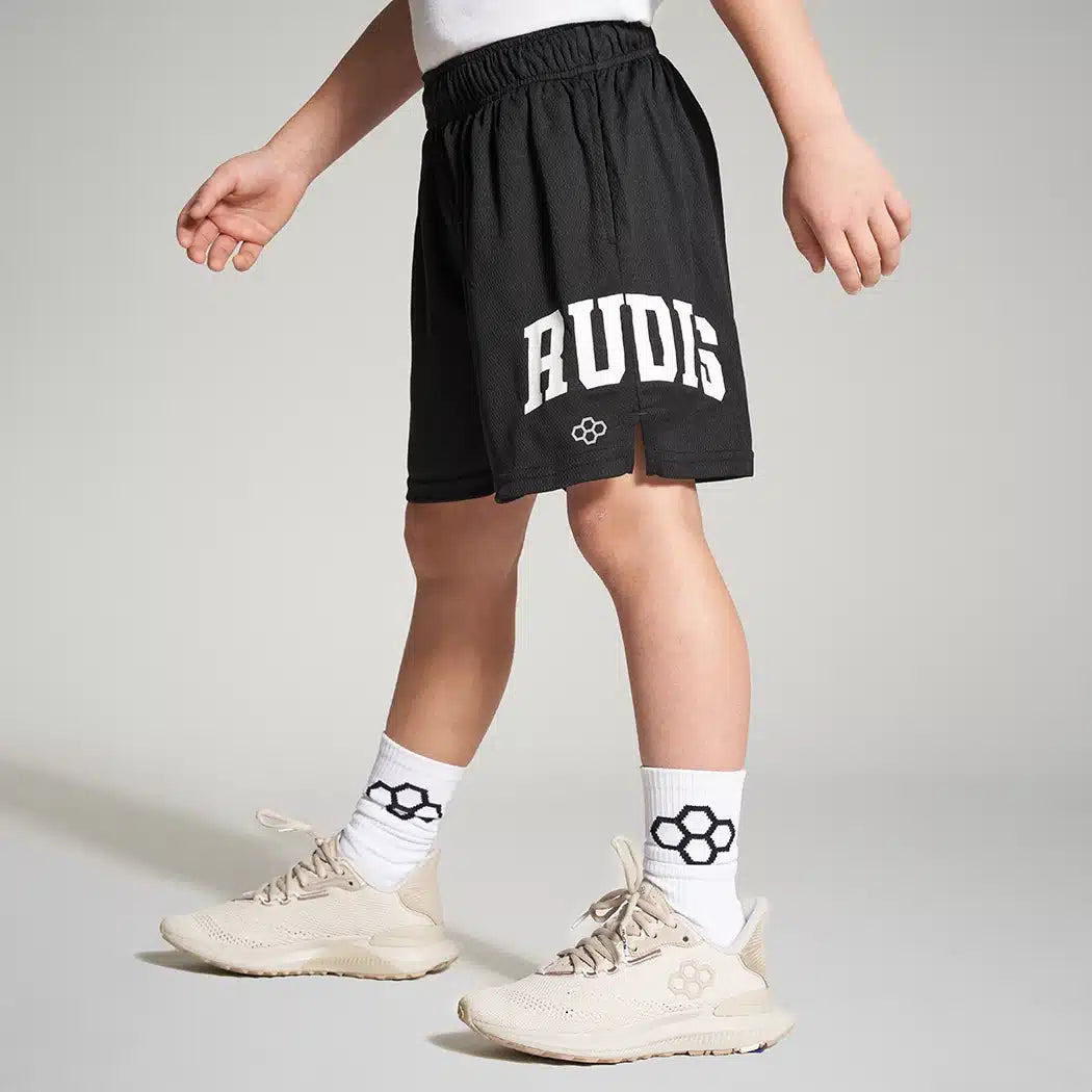 A young child wearing black athletic shorts with the word RUDIS printed on them paired with light-colored sneakers and patterned socks