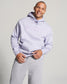 A man wearing a light lavender hoodie with RUDIS printed on the chest paired with gray sweatpants smiles confidently against a soft background