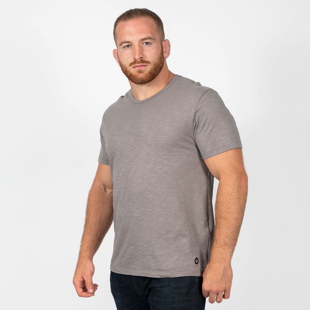 A muscular man stands confidently in a light gray t-shirt and dark jeans showcasing a casual yet stylish look against a plain white background