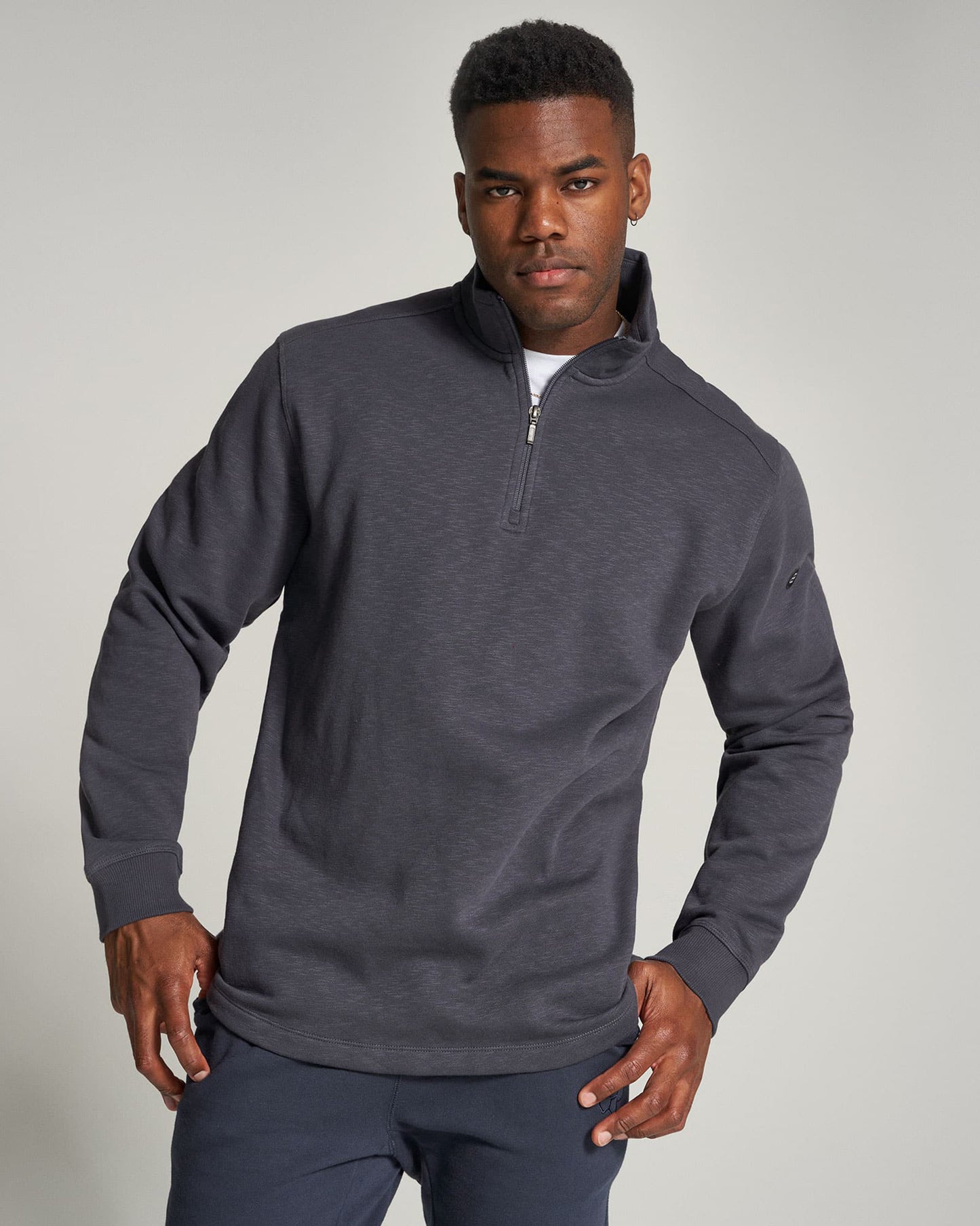 A young man wearing a dark gray zip-up pullover sweater over a white t-shirt posing confidently against a neutral background