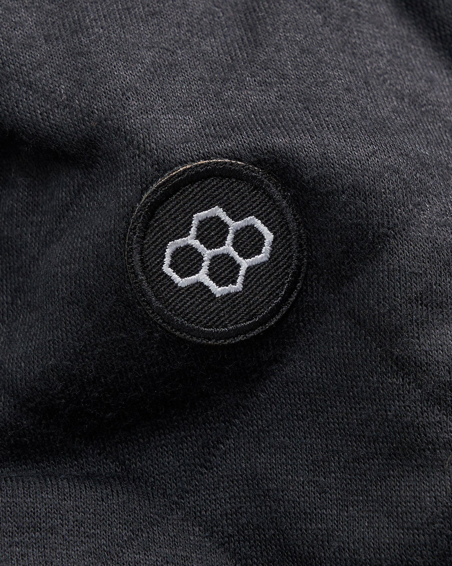 A close-up view of a black fabric displaying a circular embroidered logo featuring hexagonal shapes in white