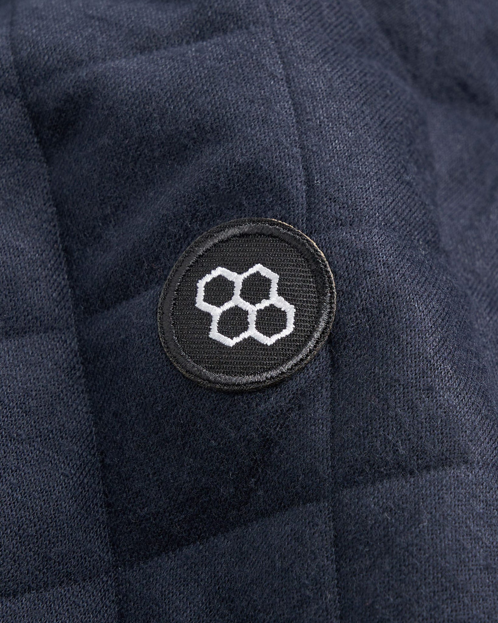 A close-up of a textured dark fabric featuring a circular black patch with a white hexagonal logo emphasizing the garments design and branding