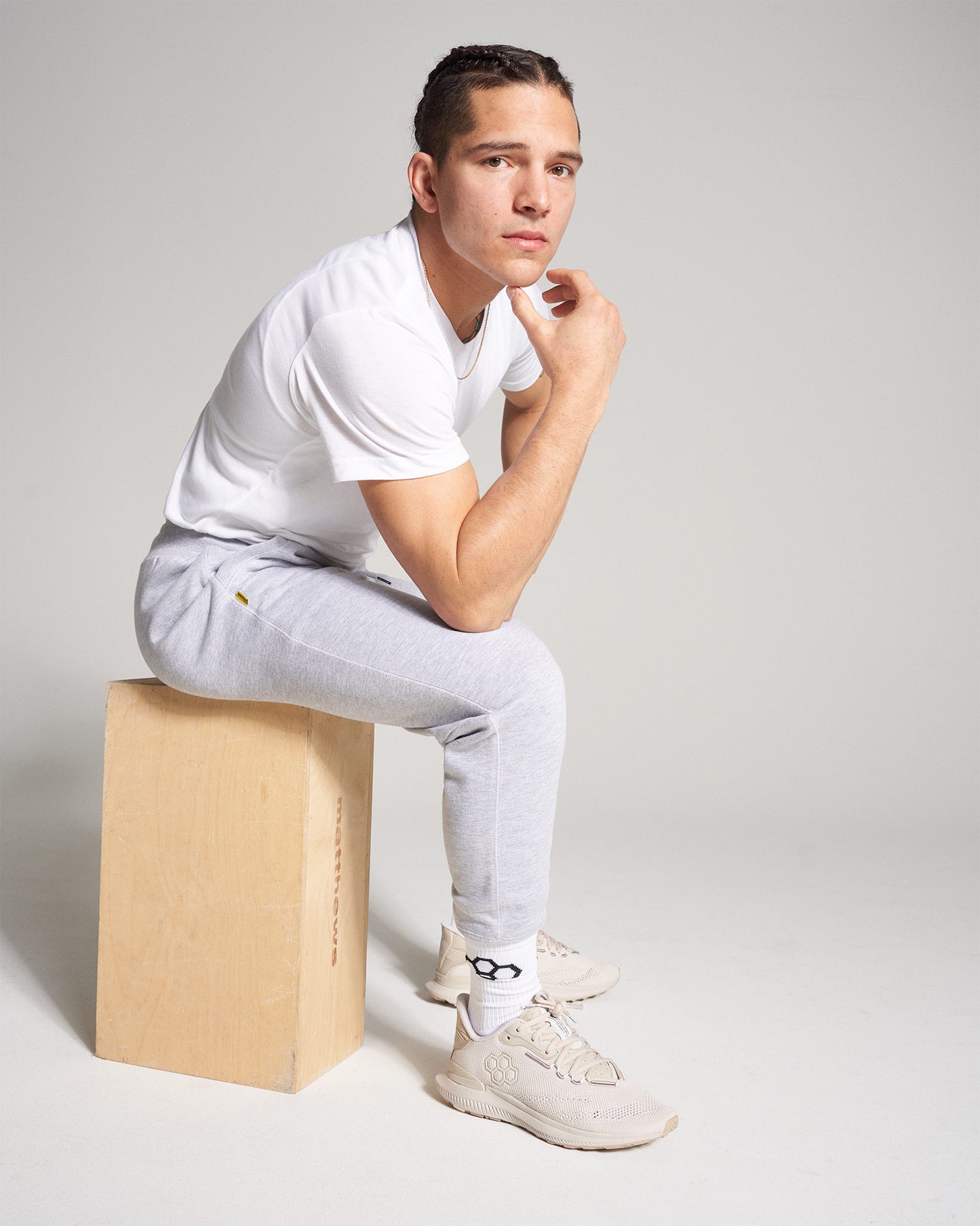 A young man with braided hair sits on a wooden box wearing a white t-shirt and gray sweatpants paired with beige sneakers and white socks projecting a contemplative mood