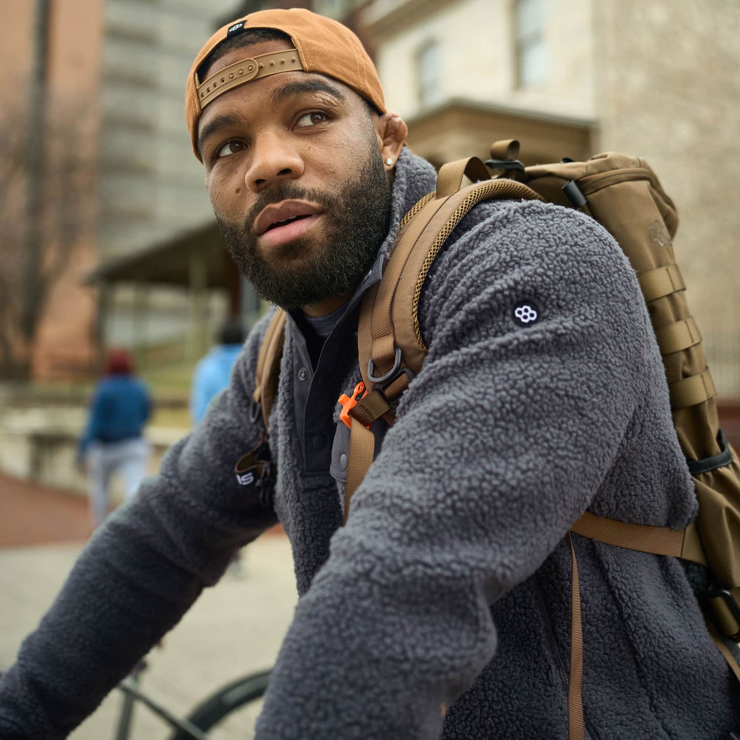 A man with a beard wearing a gray fleece jacket and a cap sitting on a bicycle with a backpack set against an urban backdrop