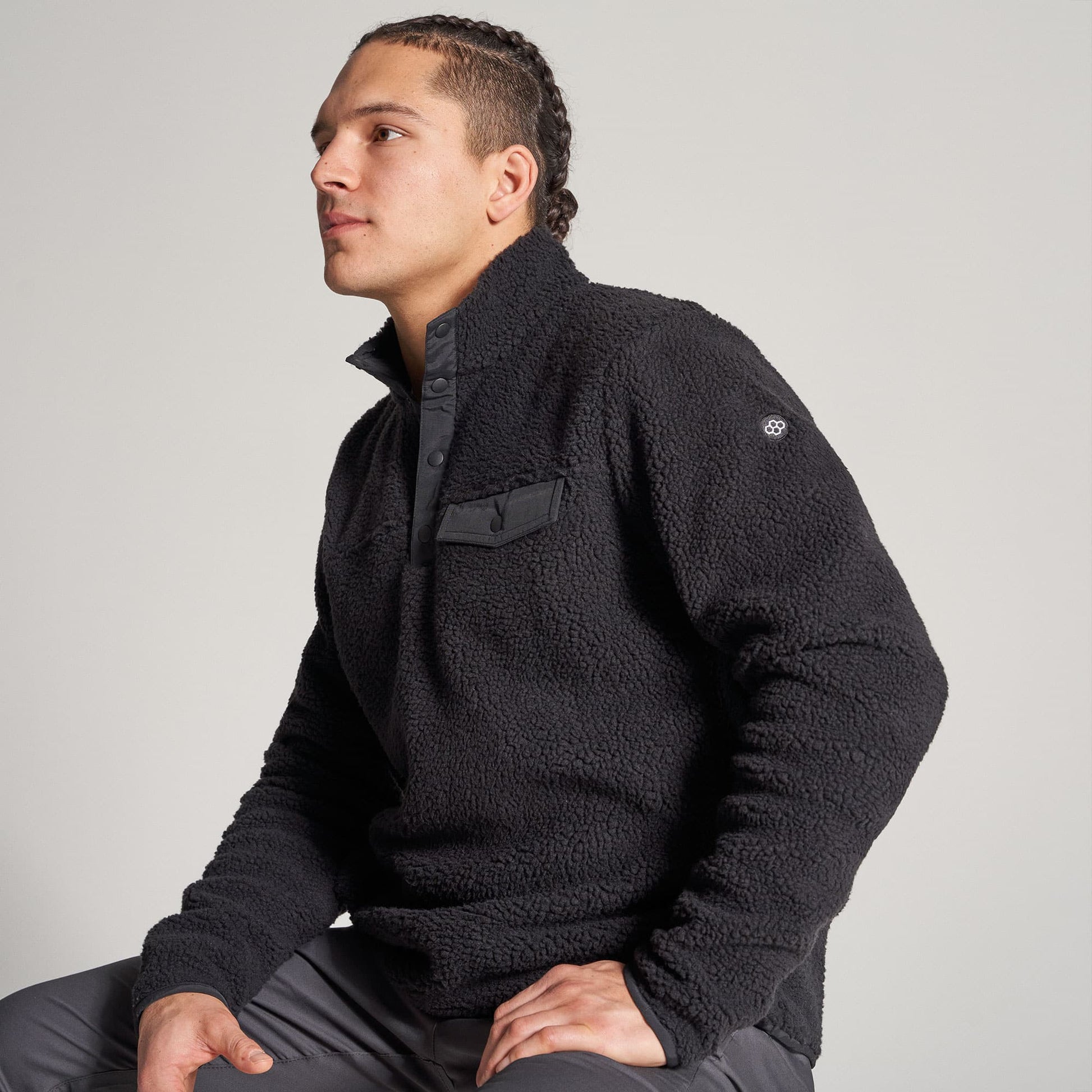 A young man is seated wearing a black fleece pullover with a half-buttoned collar and a discreet pocket showcasing a stylish and comfortable look